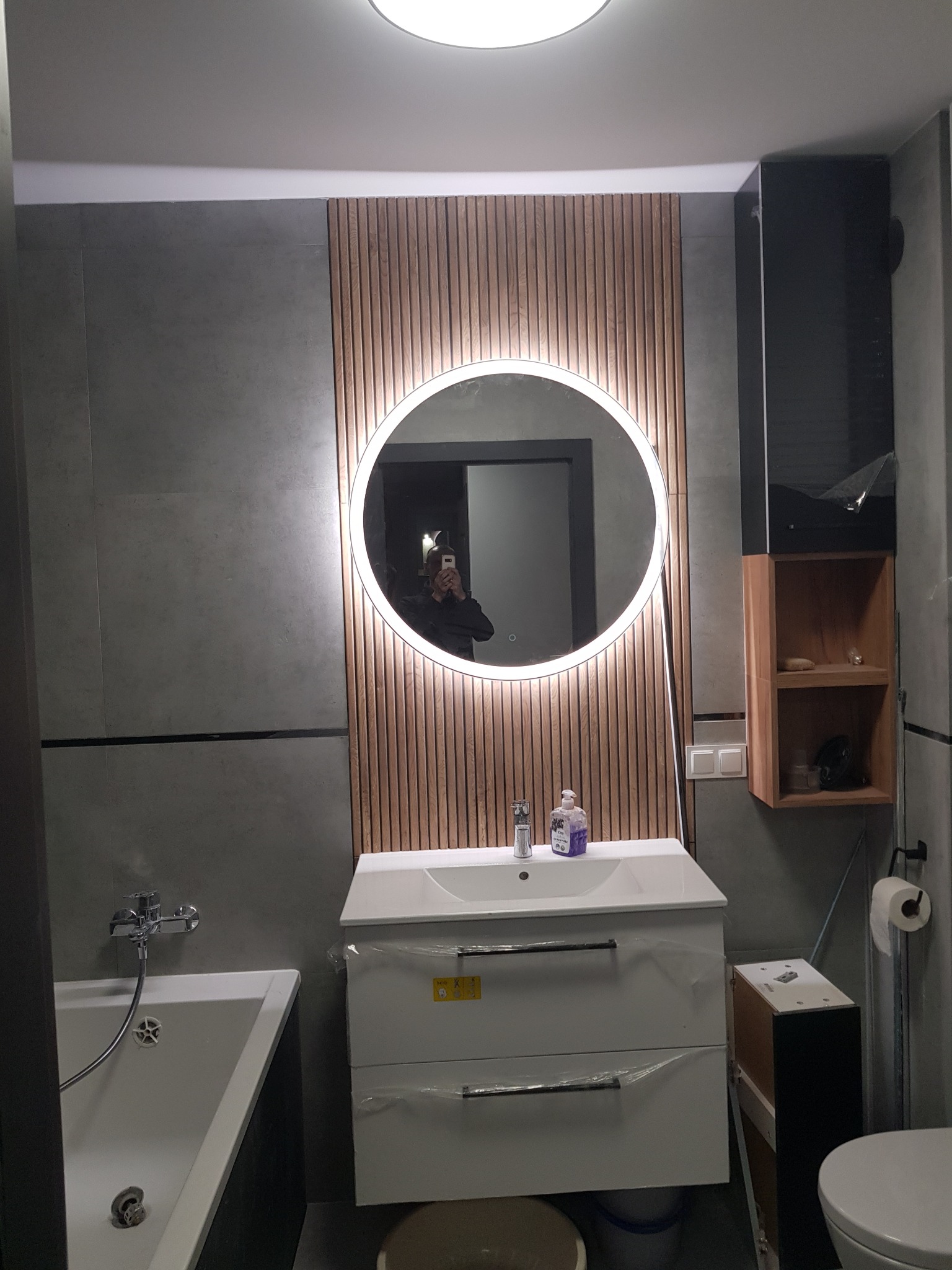 Nowoczesna łazienka z okrągłym lustrem podświetlanym LED, umywalką z szafką w folii ochronnej, drewnianymi panelami dekoracyjnymi i szarą płytką.