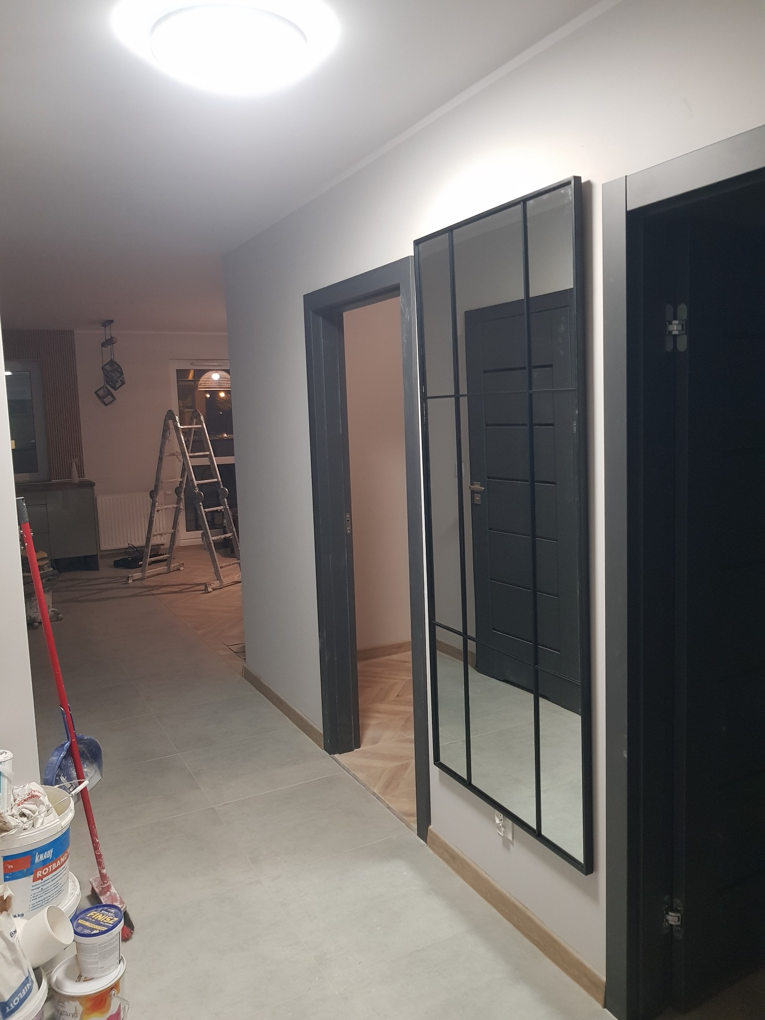 Korytarz w trakcie wykańczania: szare ściany, płytki podłogowe, czarne drzwi i lustro w metalowej ramie, widoczne wiadro z farbą i drabina w tle.