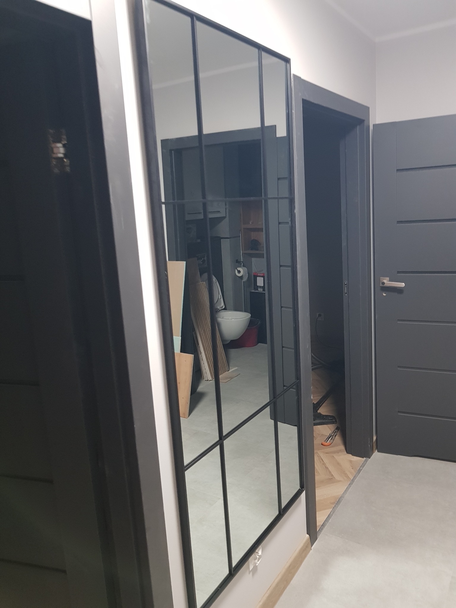 Szklana ścianka działowa z czarnymi profilami w trakcie remontu łazienki, widoczne narzędzia i materiały budowlane w odbiciu lustrzanym, surowy stan wykończenia.