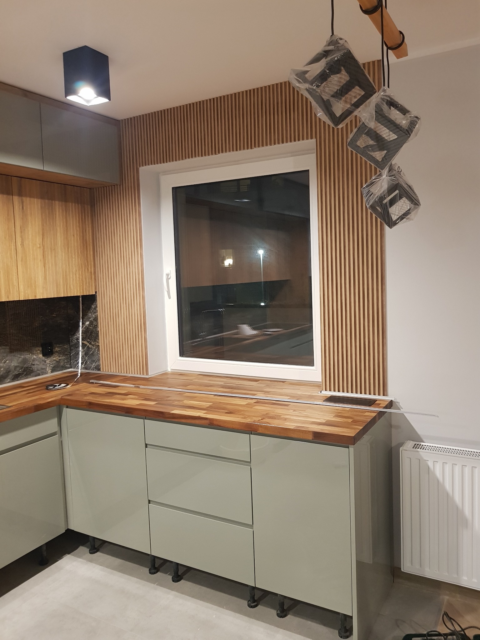 Nowoczesna kuchnia z drewnianym blatem, szarymi szafkami i dekoracyjnymi lamelami ściennymi za oknem, wiszące lampy w folii ochronnej.