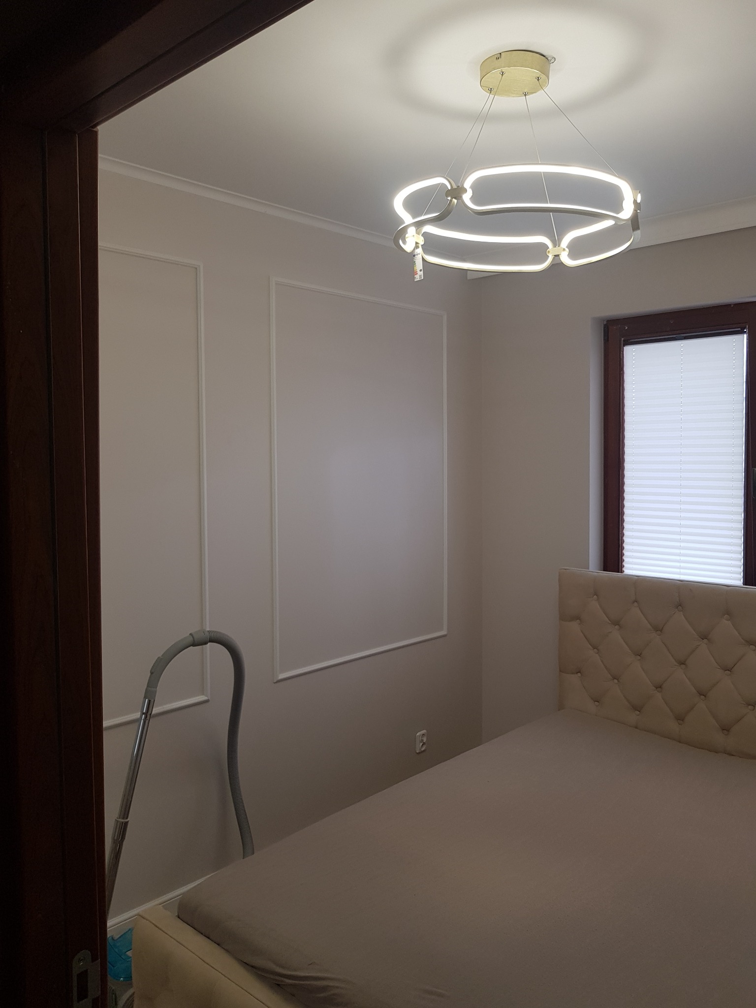 Jasna sypialnia z dekoracyjnymi panelami ściennymi, nowoczesną lampą sufitową i łóżkiem z pikowanym zagłówkiem; widoczny odkurzacz przy ścianie.