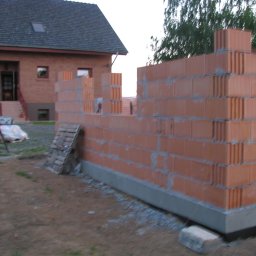 Grzegorz Naumowicz - Wznoszenie ściany z czerwonej cegły na fundamencie. Widoczny fragment domu w tle, palety i gruz. Etap budowy domu jednorodzinnego.