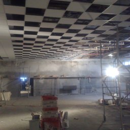 Grzegorz Naumowicz - Montaż sufitu podwieszanego kasetonowego w szachownicę. Widoczne rusztowania, worki z zaprawą i pracownik na rusztowaniu. Hala w trakcie prac wykończeniowych.