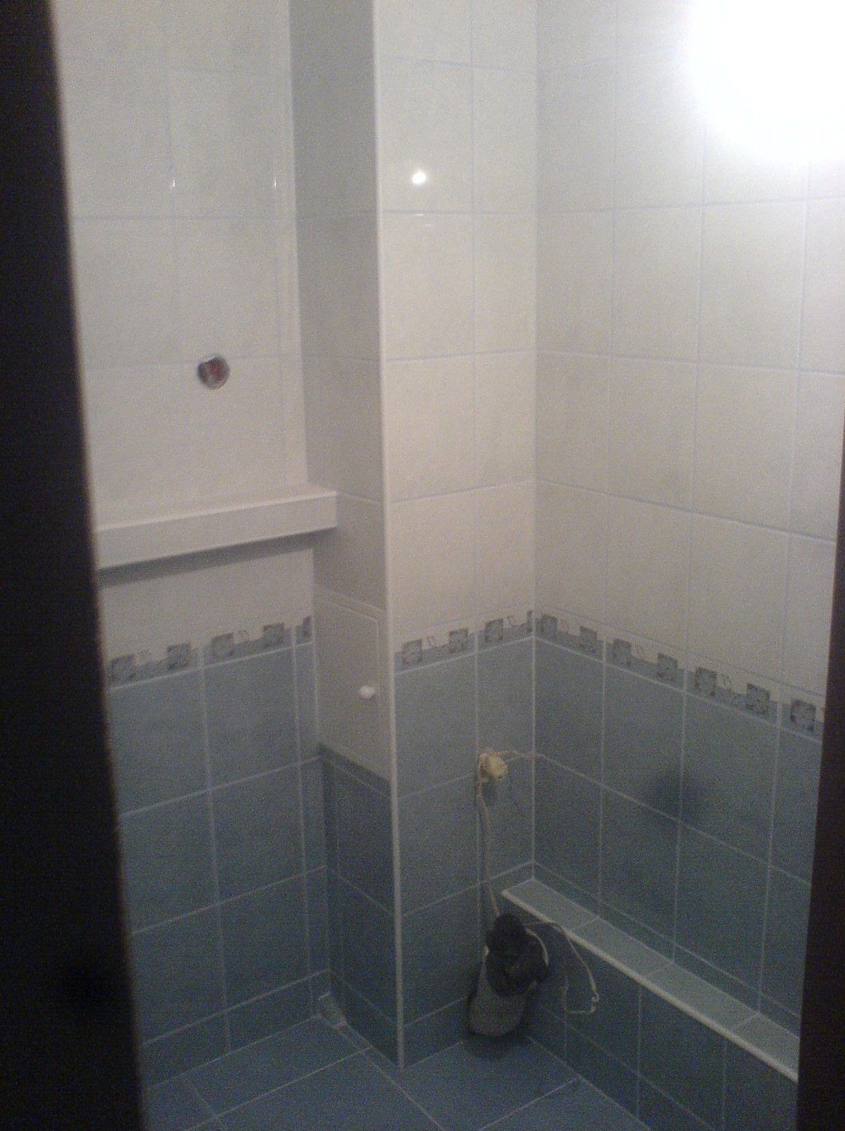 Wykończona łazienka z dwukolorowymi płytkami, gdzie dolna część jest niebieska z dekoracyjnym pasem, a górna biała. Widoczna półka i wystający kabel.