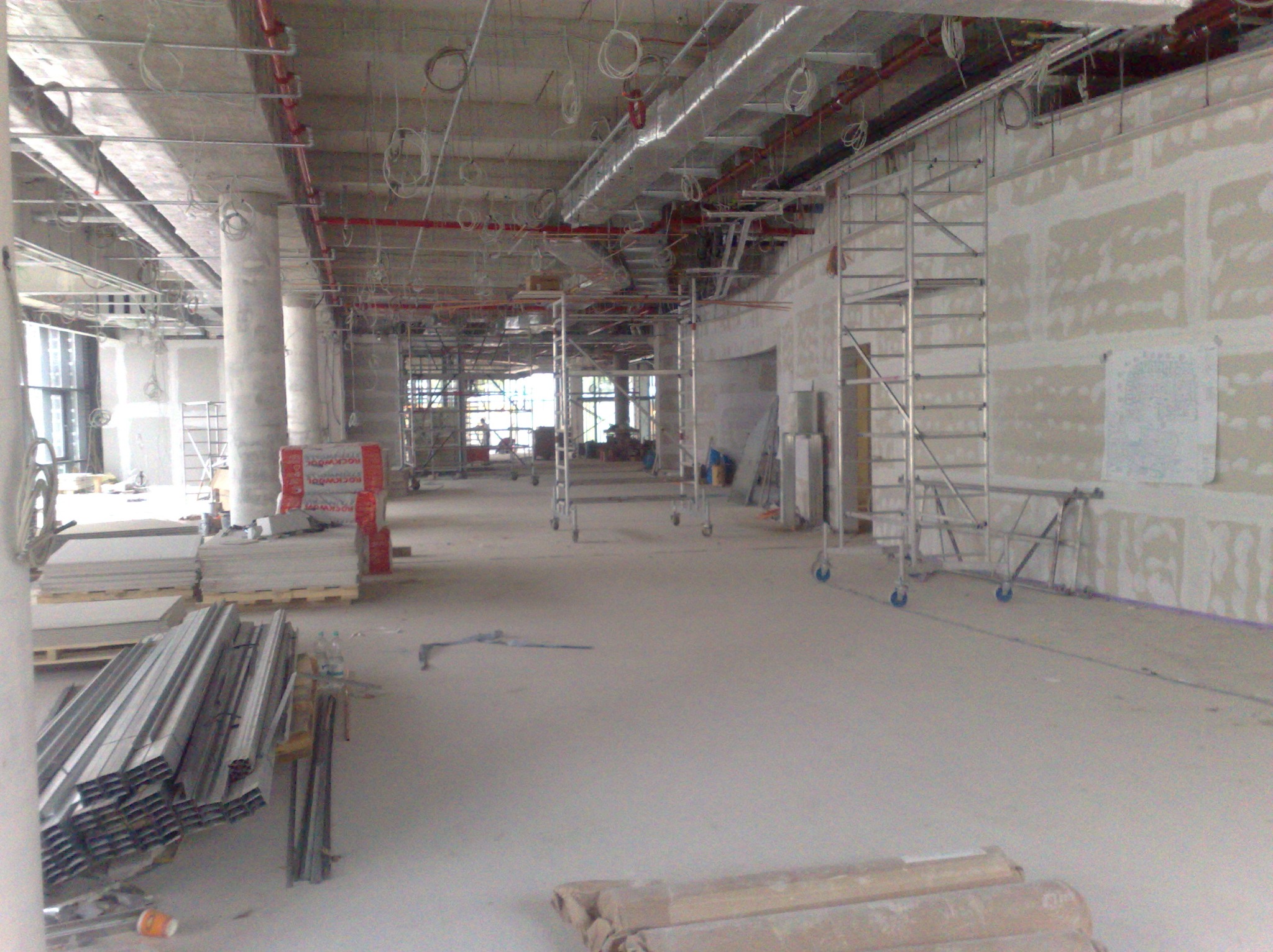 Wnętrze w trakcie prac wykończeniowych: widoczne rusztowania, płyty gipsowe, profile metalowe i instalacje pod sufitem. Surowy stan, przygotowanie do dalszych prac.