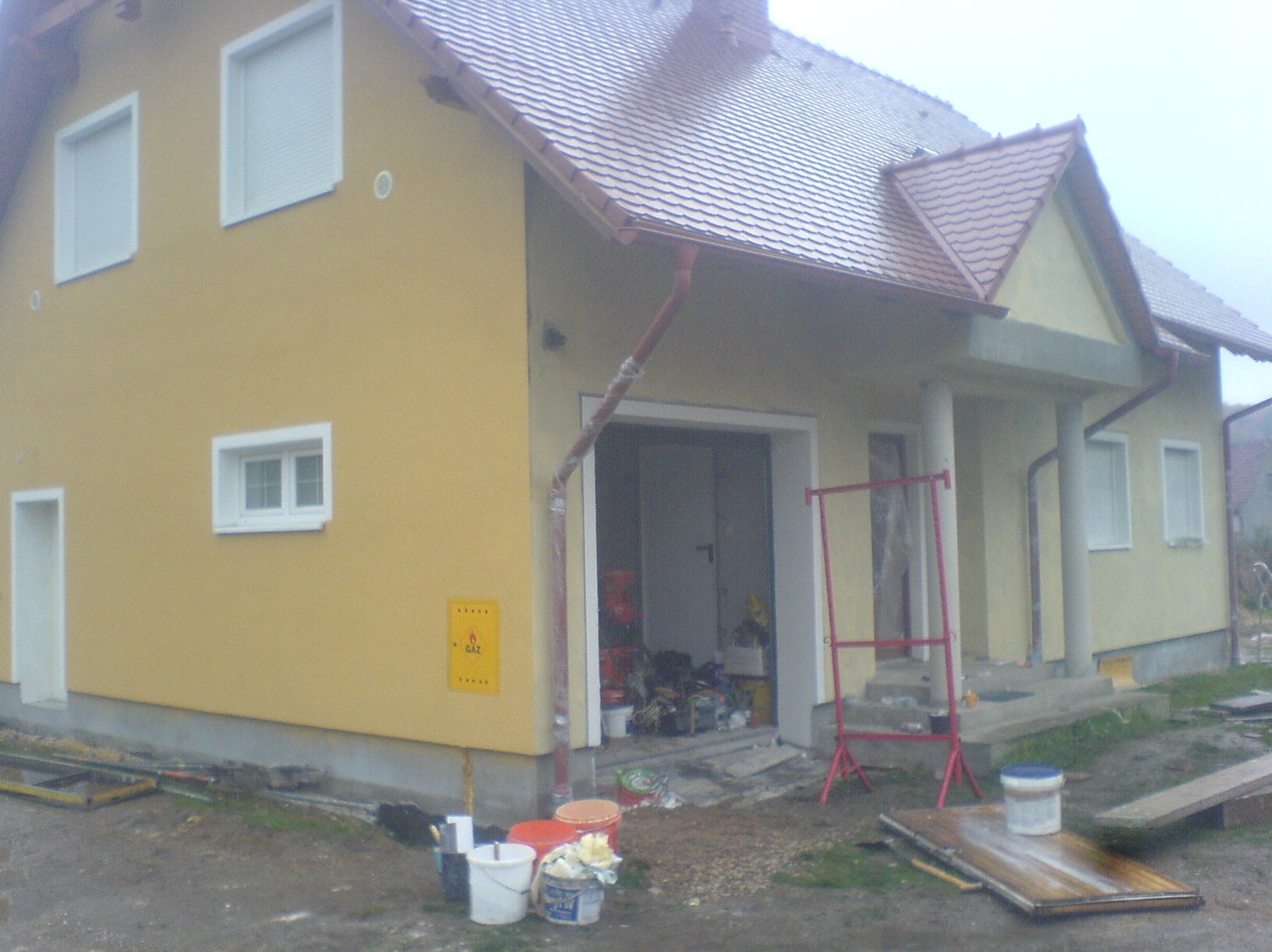 Żółty dom jednorodzinny w trakcie prac wykończeniowych. Widoczne rusztowanie, wiaderka z farbą i narzędzia. Częściowo otynkowana elewacja i nowe okna.