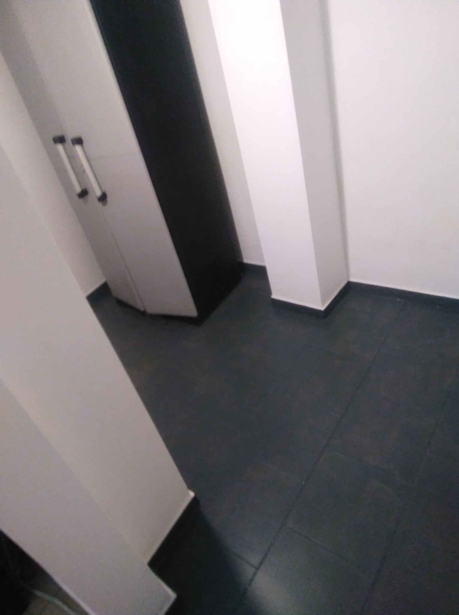 Ciemna, świeżo ułożona podłoga z płytek w pustym pomieszczeniu z białymi ścianami i kolumną. Widoczna szafa wnękowa z srebrnymi uchwytami. Ujęcie z dołu.