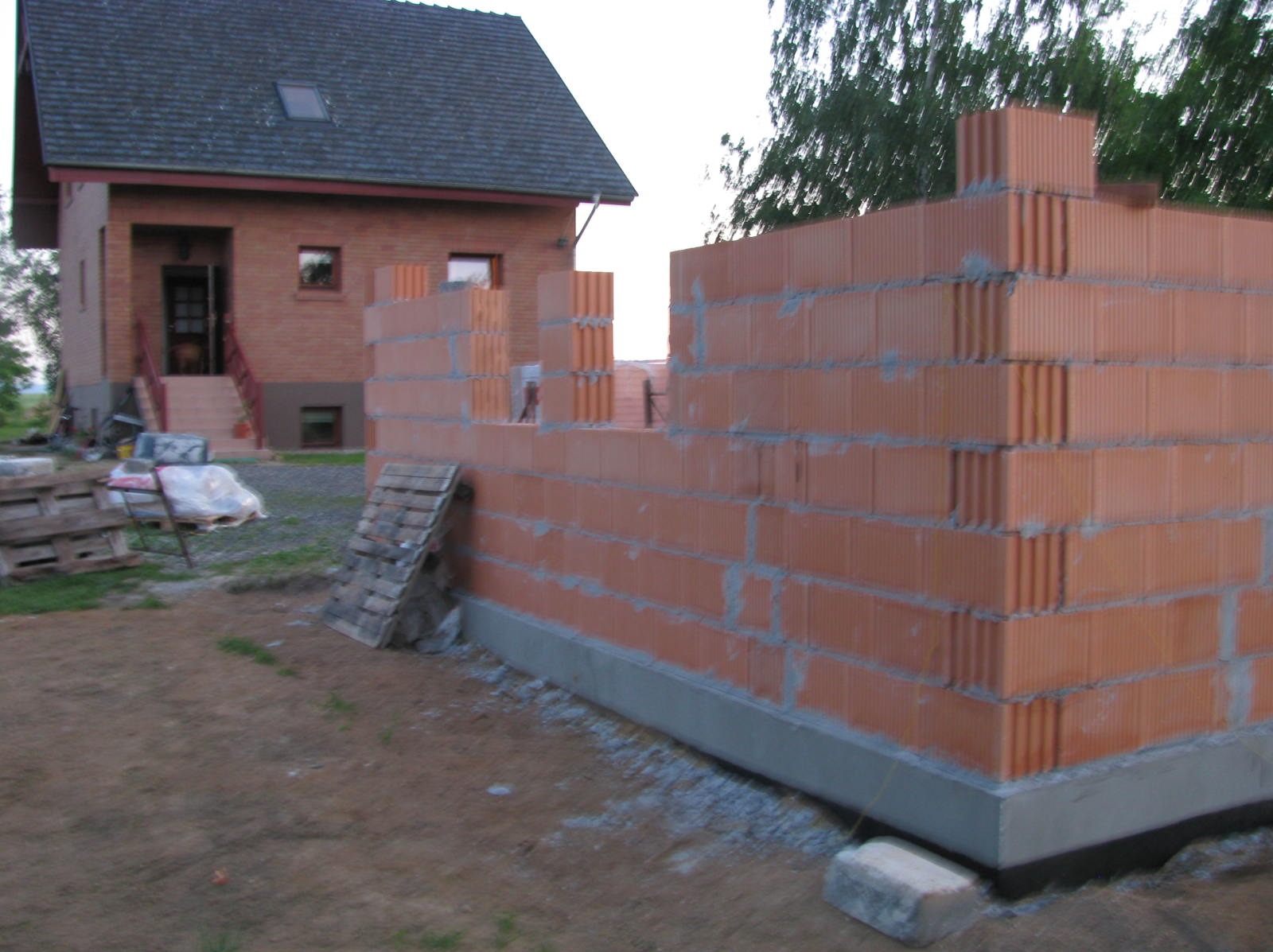 Wznoszenie ściany z czerwonej cegły na fundamencie. Widoczny fragment domu w tle, palety i gruz. Etap budowy domu jednorodzinnego.