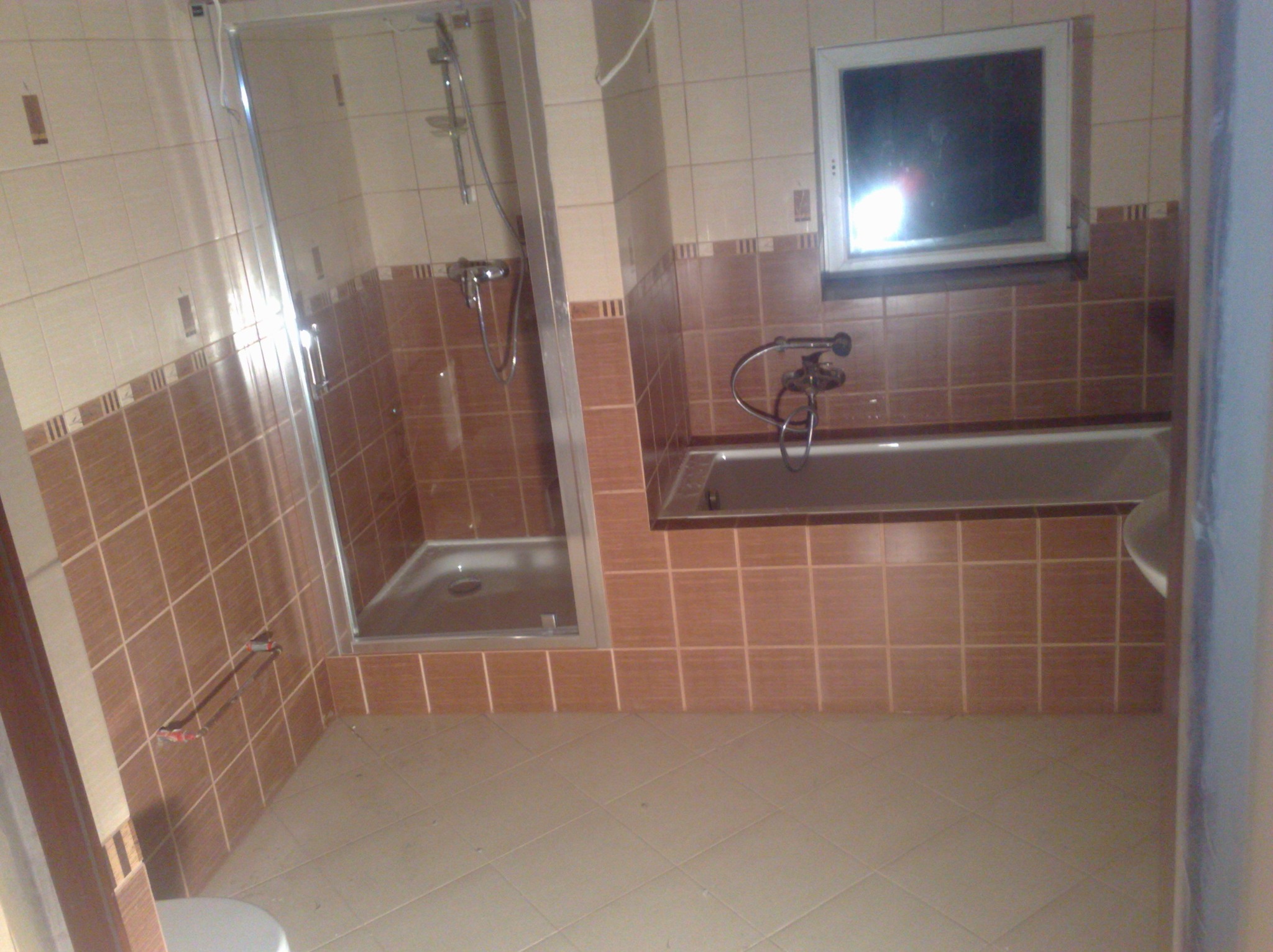 Wykończona łazienka z brązowo-beżowymi płytkami, kabiną prysznicową i wanną pod oknem. Widoczne baterie i akcesoria łazienkowe. Ukończony remont.