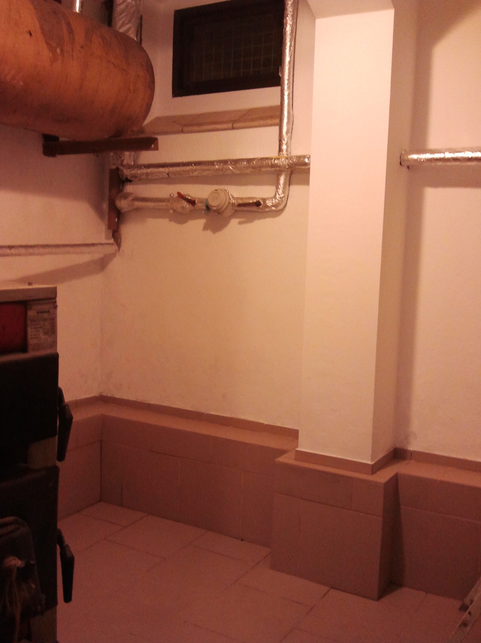 Zakończona instalacja CO w piwnicy: piec, zbiornik, rury w izolacji, brązowe płytki na podłodze i cokole. Widok na fragment kotłowni po remoncie.