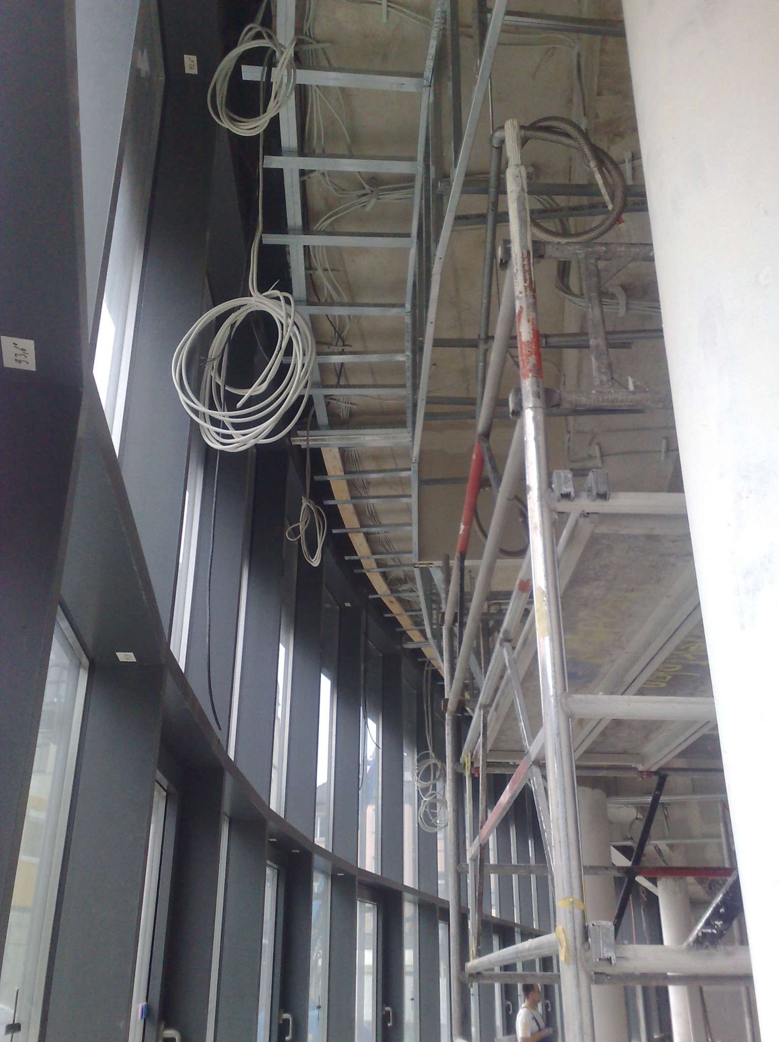 Wnętrze w trakcie prac wykończeniowych: widoczne rusztowania, instalacje elektryczne zwisające z sufitu, metalowa konstrukcja podwieszanego sufitu i okna.