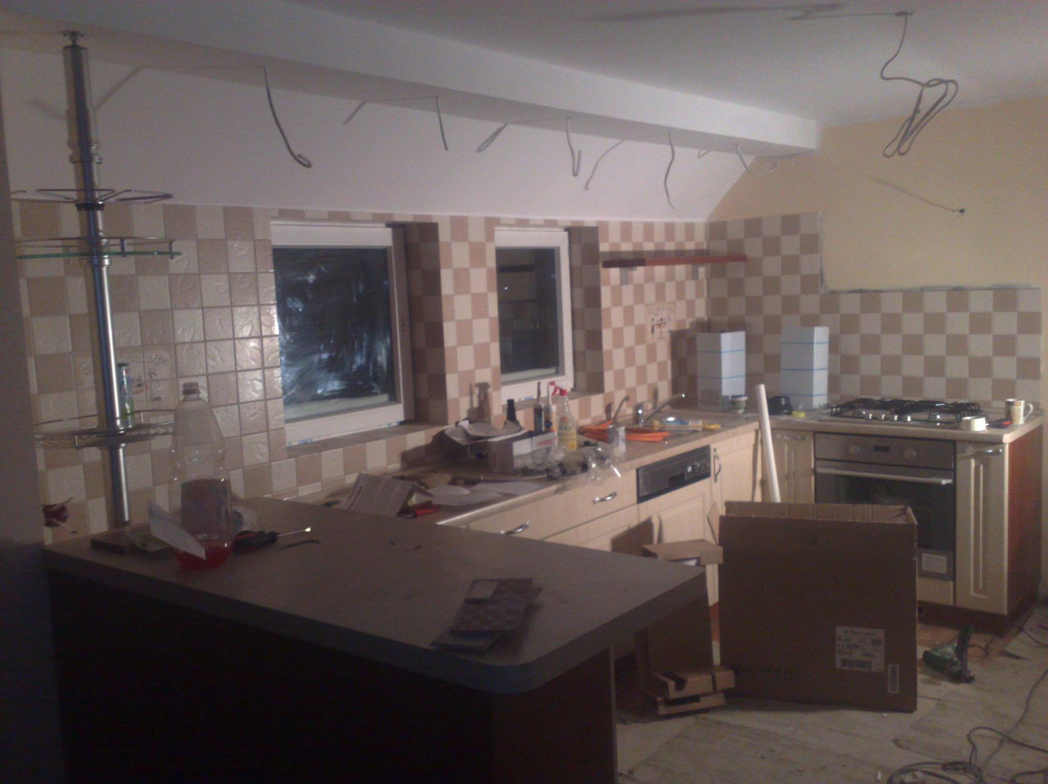 Kuchnia w trakcie remontu: częściowo ułożone płytki w szachownicę, wiszące kable, sprzęt AGD i narzędzia. Widok na wnętrze z elementami wykończeniowymi.