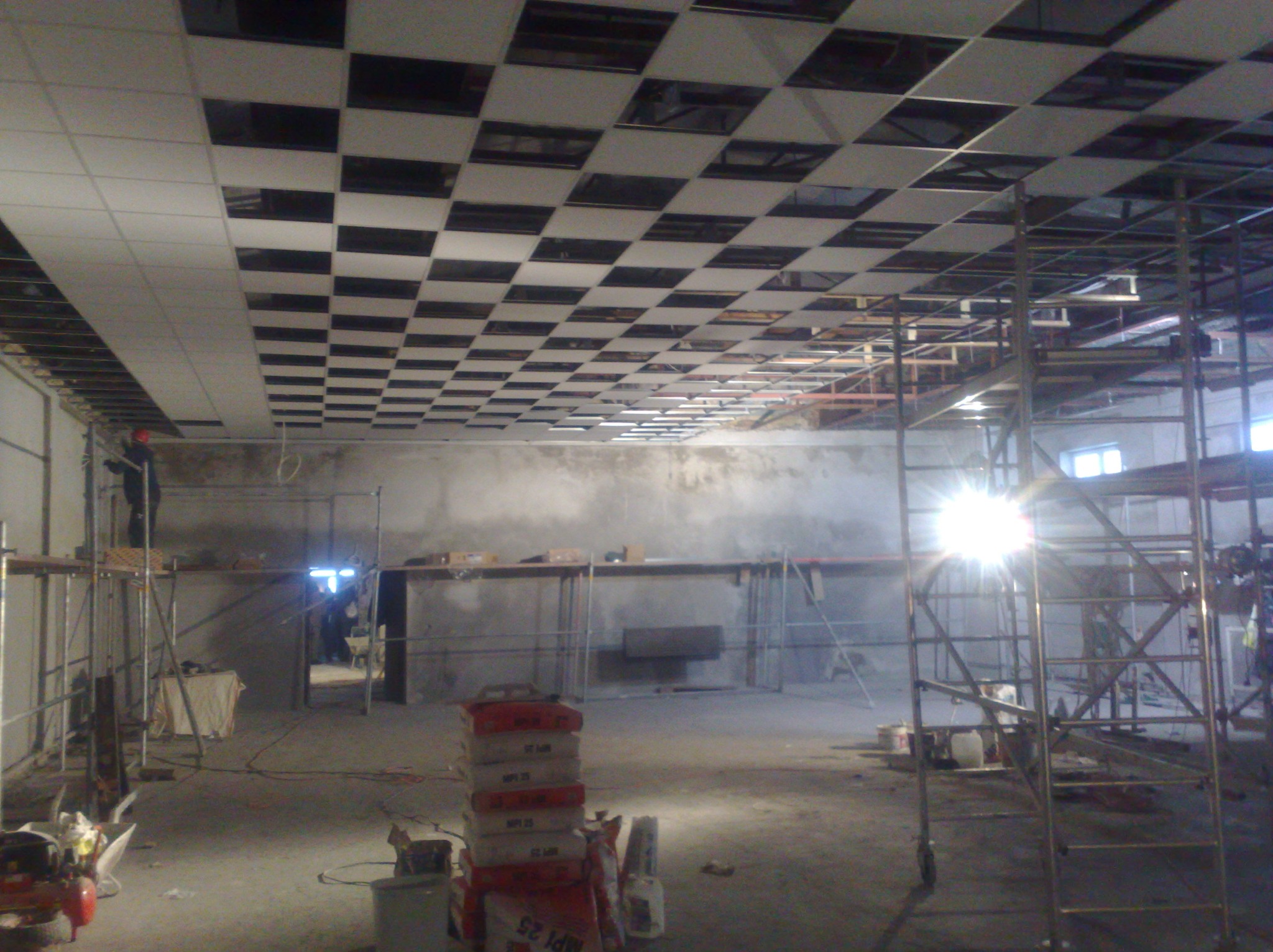 Montaż sufitu podwieszanego kasetonowego w szachownicę. Widoczne rusztowania, worki z zaprawą i pracownik na rusztowaniu. Hala w trakcie prac wykończeniowych.
