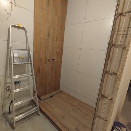 Mark-Art-Rem Marek Piwoński - Wykończenie łazienki w trakcie: ułożone płytki imitujące drewno na podłodze i ścianie prysznica, białe płytki na ścianach, drabina aluminiowa, surowy mur z boku.