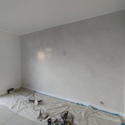 Mark-Art-Rem Marek Piwoński - Pokój po nałożeniu dekoracyjnej, szarej gładzi na ścianie. Podłoga zabezpieczona folią malarską, widoczne narzędzia pracy. Minimalistyczne wnętrze z dużym oknem.