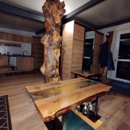 Mark-Art-Rem Marek Piwoński - Drewniane wnętrze z unikalnym stołem z żywicą i pniem drzewa jako elementem konstrukcyjnym. Widoczna kuchnia i szafa w tle. Ciepłe oświetlenie.