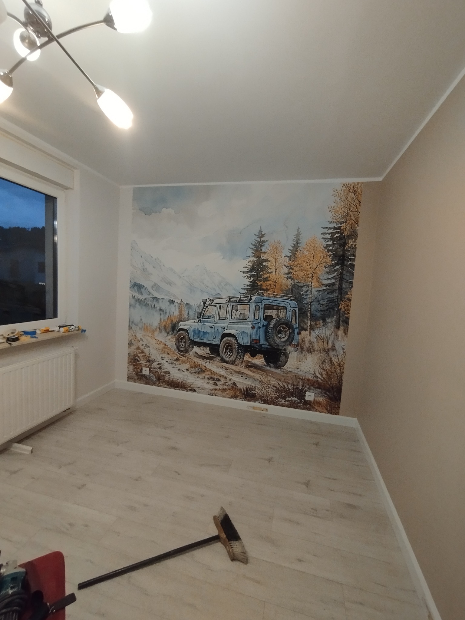 Pokój po malowaniu z fototapetą przedstawiającą terenowy samochód na tle górskiego krajobrazu. Na podłodze leżą narzędzia i szczotka.