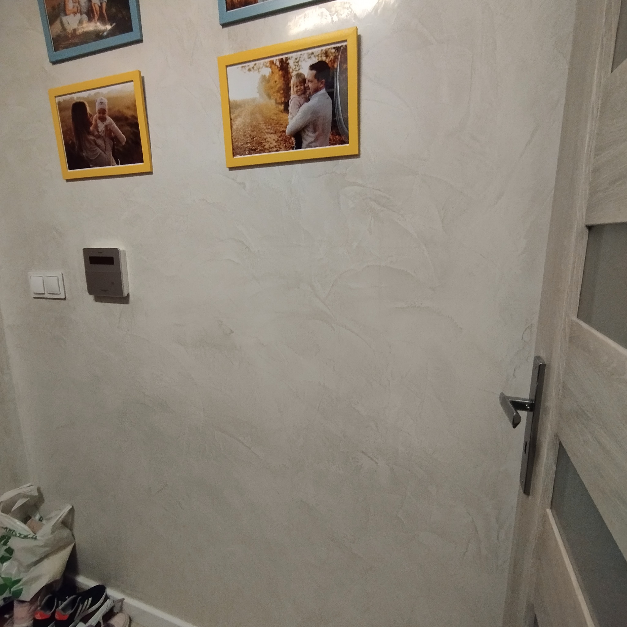 Ściana z dekoracyjną, szarą strukturą, ozdobiona ramkami ze zdjęciami rodzinnymi. Widoczny fragment drzwi i włącznik światła.
