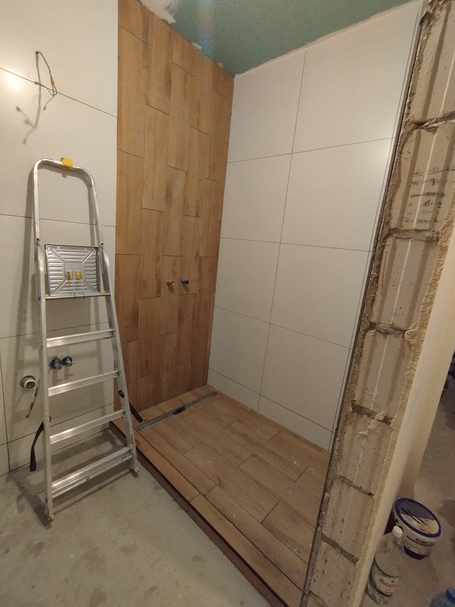 Wykończenie łazienki w trakcie: ułożone płytki imitujące drewno na podłodze i ścianie prysznica, białe płytki na ścianach, drabina aluminiowa, surowy mur z boku.