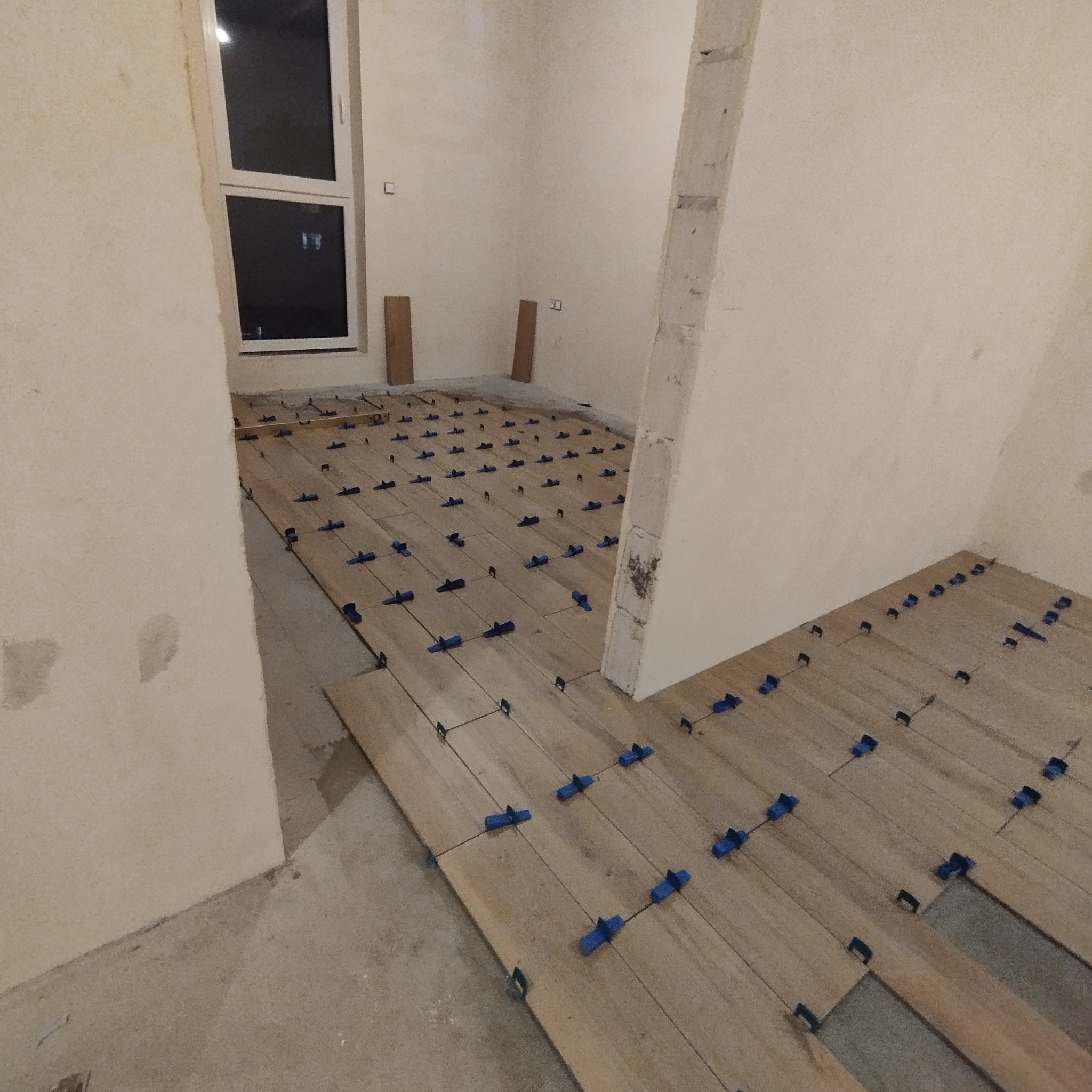 Układanie drewnianych paneli podłogowych z użyciem niebieskich klipsów poziomujących w surowym wnętrzu. Widoczne fragmenty nieułożonej podłogi i okno.