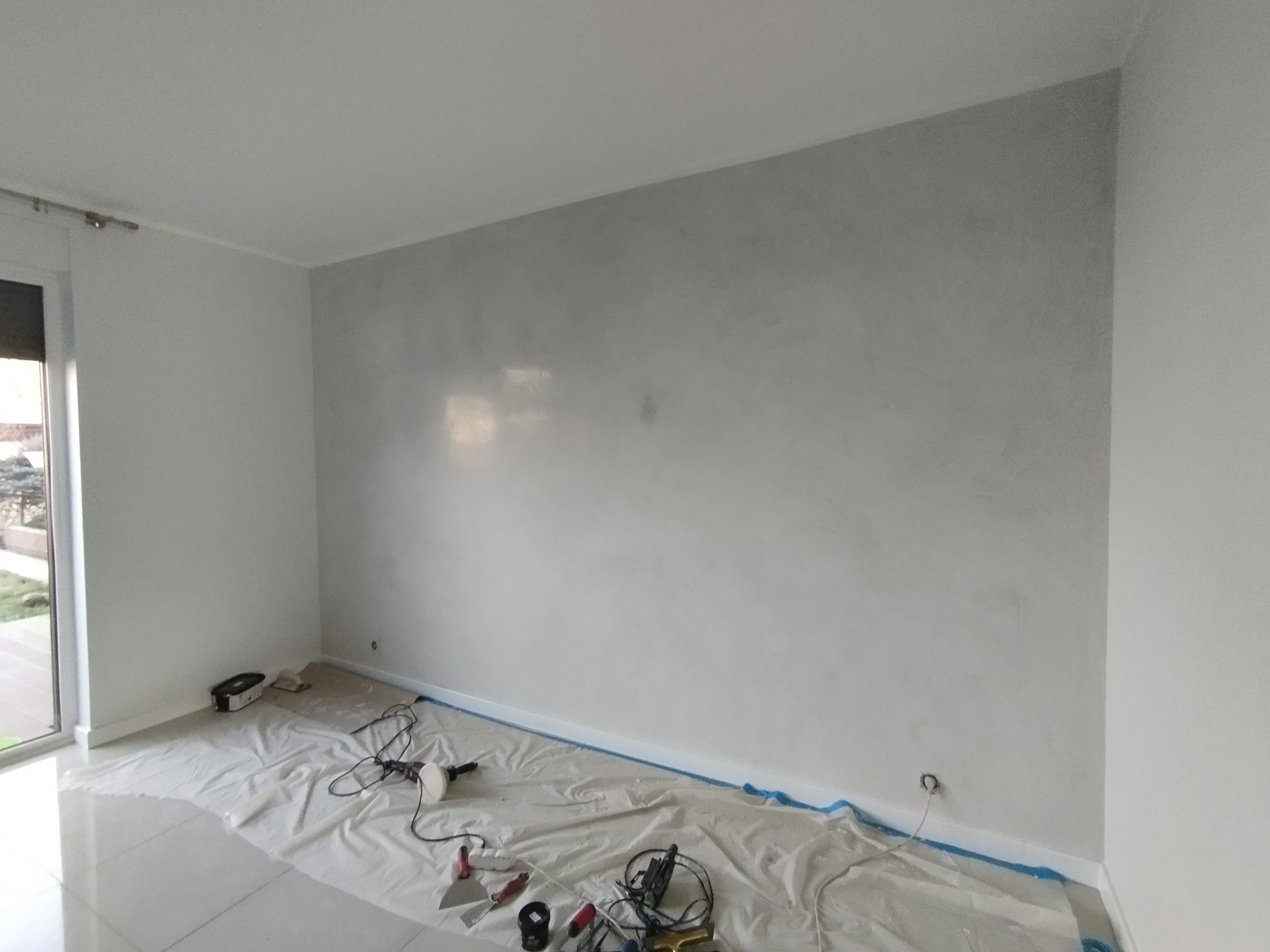 Pokój po nałożeniu dekoracyjnej, szarej gładzi na ścianie. Podłoga zabezpieczona folią malarską, widoczne narzędzia pracy. Minimalistyczne wnętrze z dużym oknem.