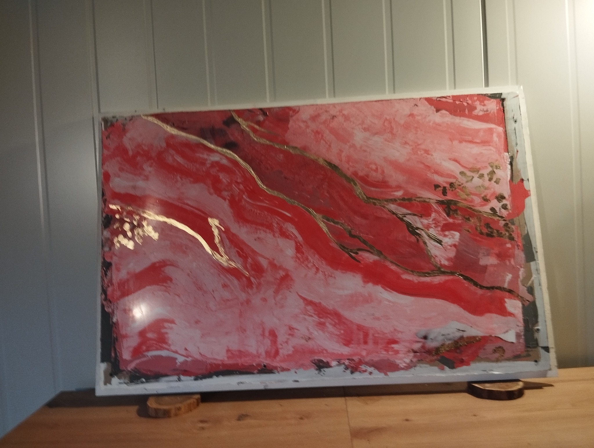 Abstrakcyjny obraz w odcieniach czerwieni i bieli z akcentami złota, umieszczony na drewnianym blacie, oparty o ścianę z paneli. Widoczne nieregularne pociągnięcia pędzla.