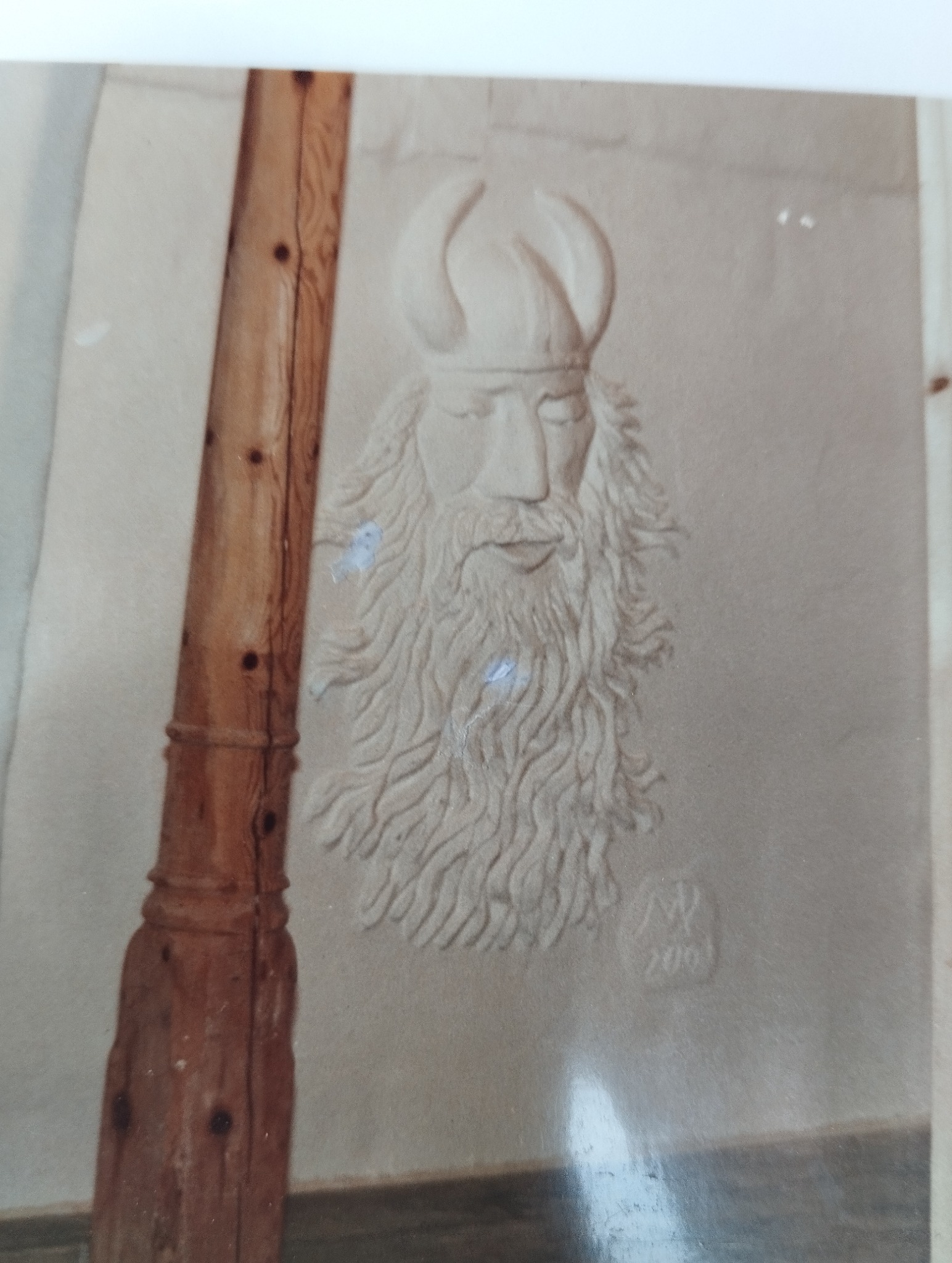 Ręcznie wykonana płaskorzeźba na ścianie przedstawiająca brodatego mężczyznę w hełmie z rogami, obok drewniany element konstrukcyjny. Sygnatura w prawym dolnym rogu.