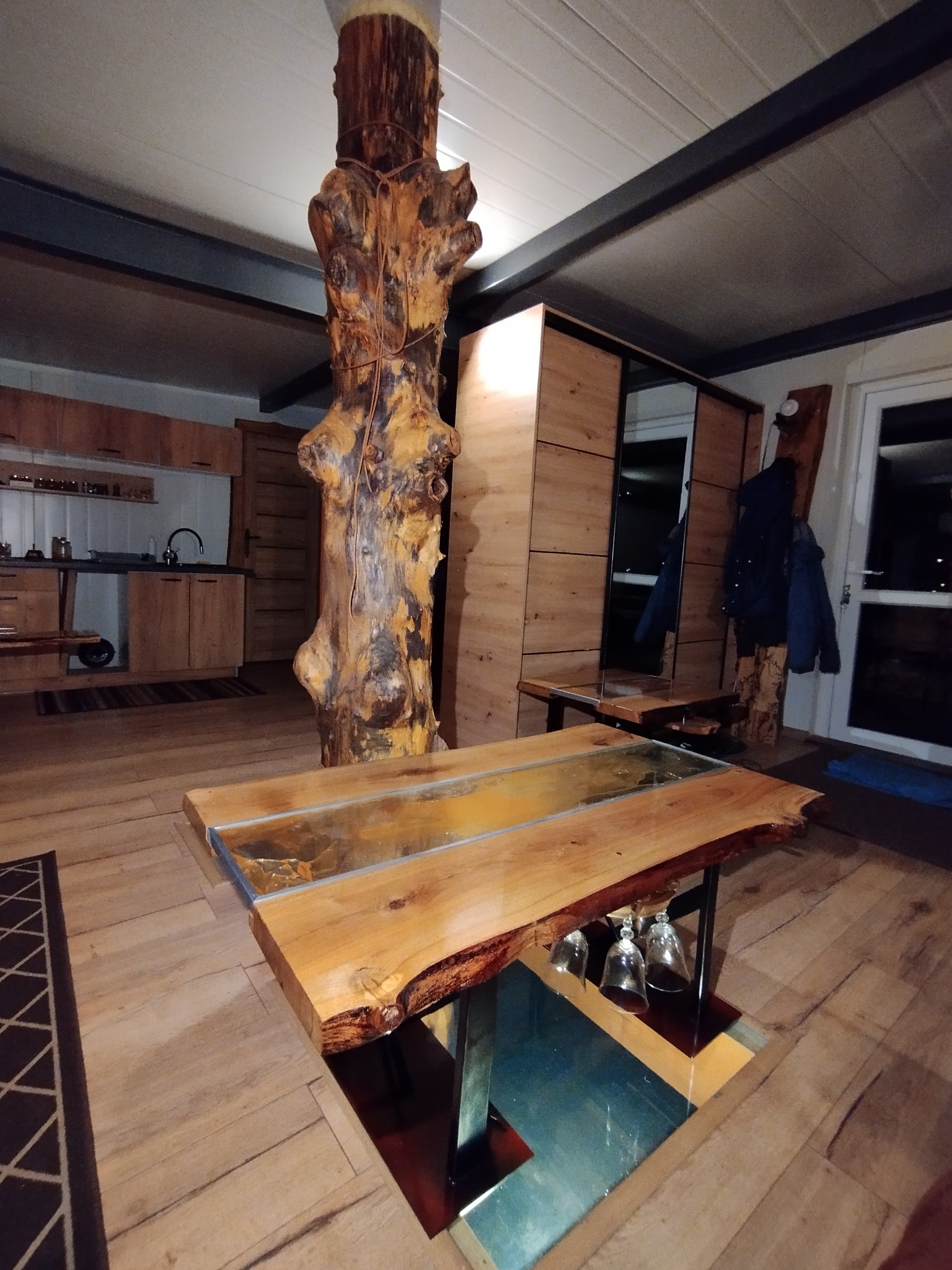 Drewniane wnętrze z unikalnym stołem z żywicą i pniem drzewa jako elementem konstrukcyjnym. Widoczna kuchnia i szafa w tle. Ciepłe oświetlenie.