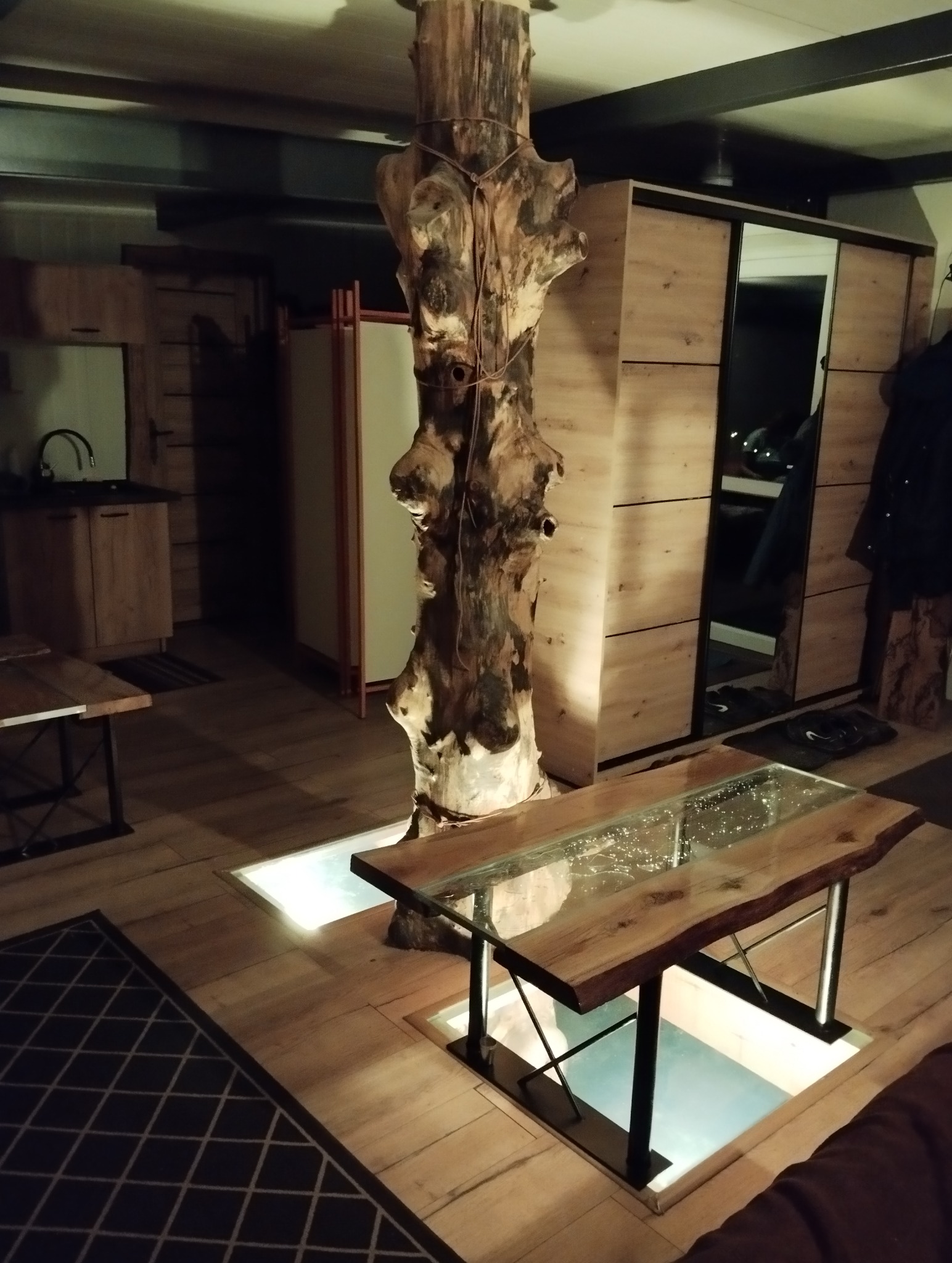 Nowoczesne wnętrze z drewnianym słupem i podświetlaną podłogą. Stół z blatem z żywicy epoksydowej. Szafa przesuwna z lustrem w tle. Unikalny design.