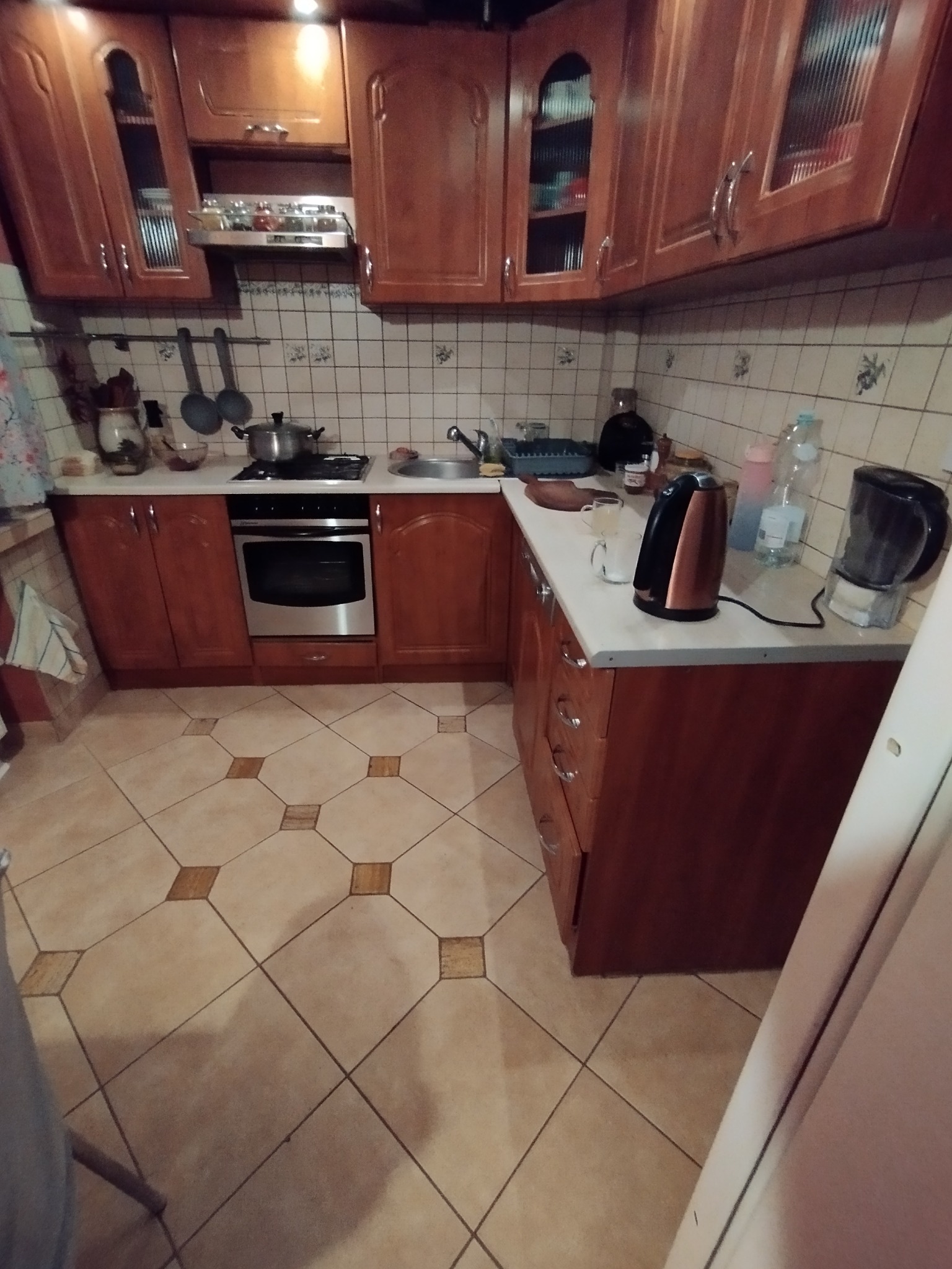 Ukończona kuchnia z brązowymi szafkami, beżowymi płytkami podłogowymi w romby i sprzętami AGD na blacie. Widok na zabudowę kuchenną.