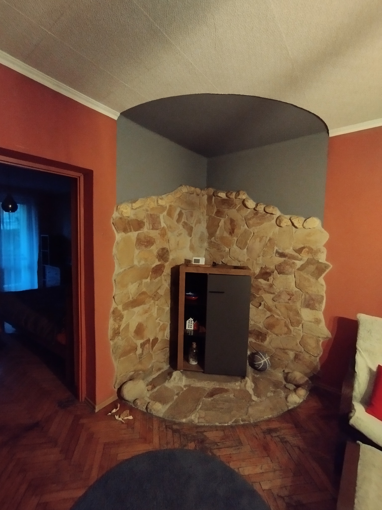 Narożna ściana z kamienia z wbudowaną szafką w salonie. Widoczna podłoga z parkietu i fragment sofy. Nietypowe wykończenie wnętrza.