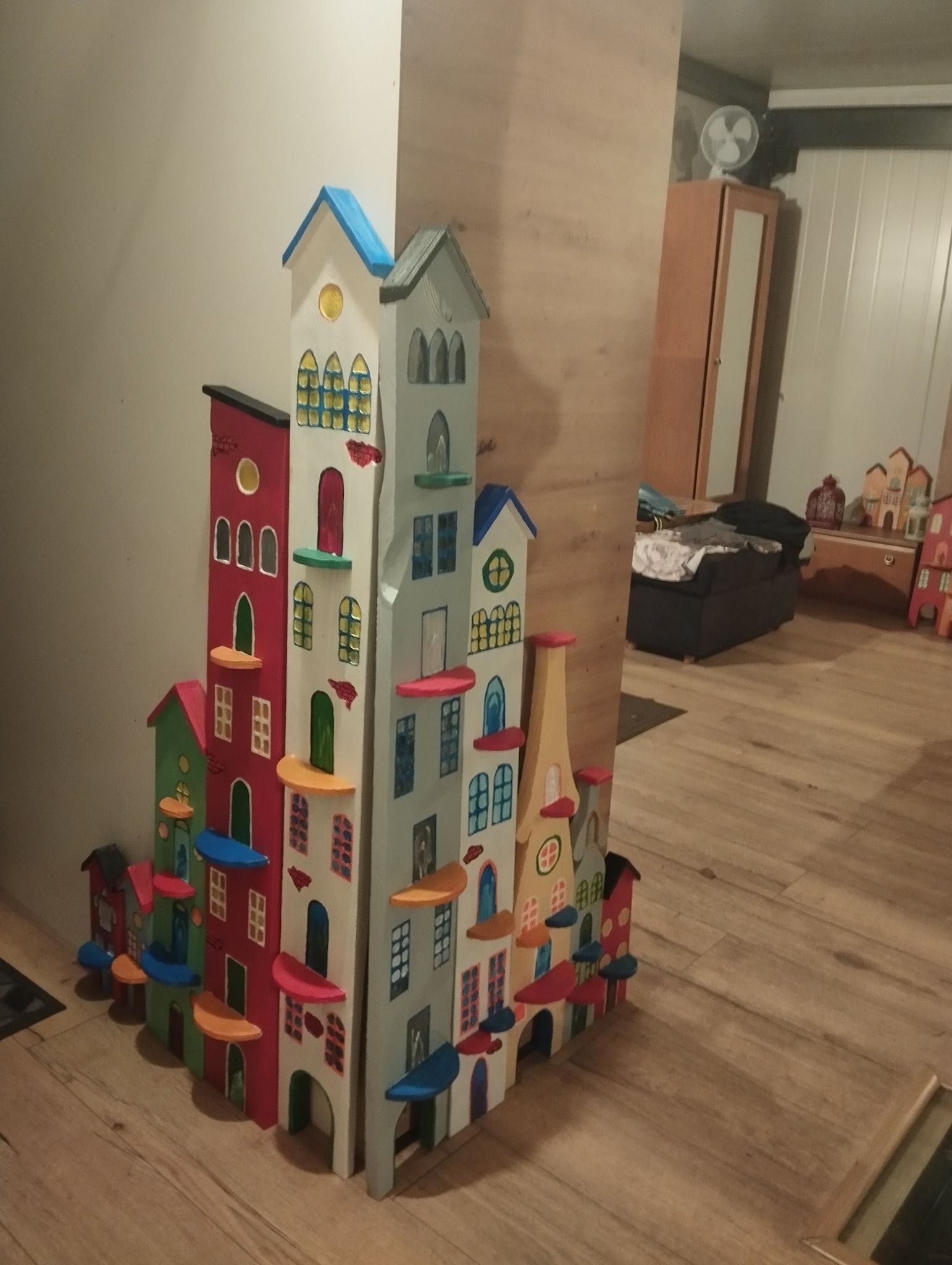 Kolorowa, drewniana dekoracja w kształcie kamieniczek, ustawiona na podłodze w pokoju. Ręcznie malowane detale, różne wysokości budynków, ciepłe oświetlenie wnętrza.