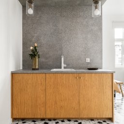 STUDIO WNĘTRZ KAROLINA PIWOWARCZYK - Nowoczesna łazienka z drewnianą szafką pod umywalką, szarym blatem i mozaikową podłogą w czarno-białe wzory. Na blacie wazon z kwiatami i czarna miseczka. Dwie szklane lampy wiszą nad blatem.