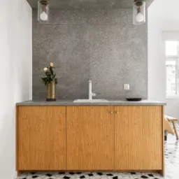 Nowoczesna łazienka z drewnianą szafką pod umywalką, szarym blatem i mozaikową podłogą w czarno-białe wzory. Na blacie wazon z kwiatami i czarna miseczka. Dwie szklane lampy wiszą nad blatem.