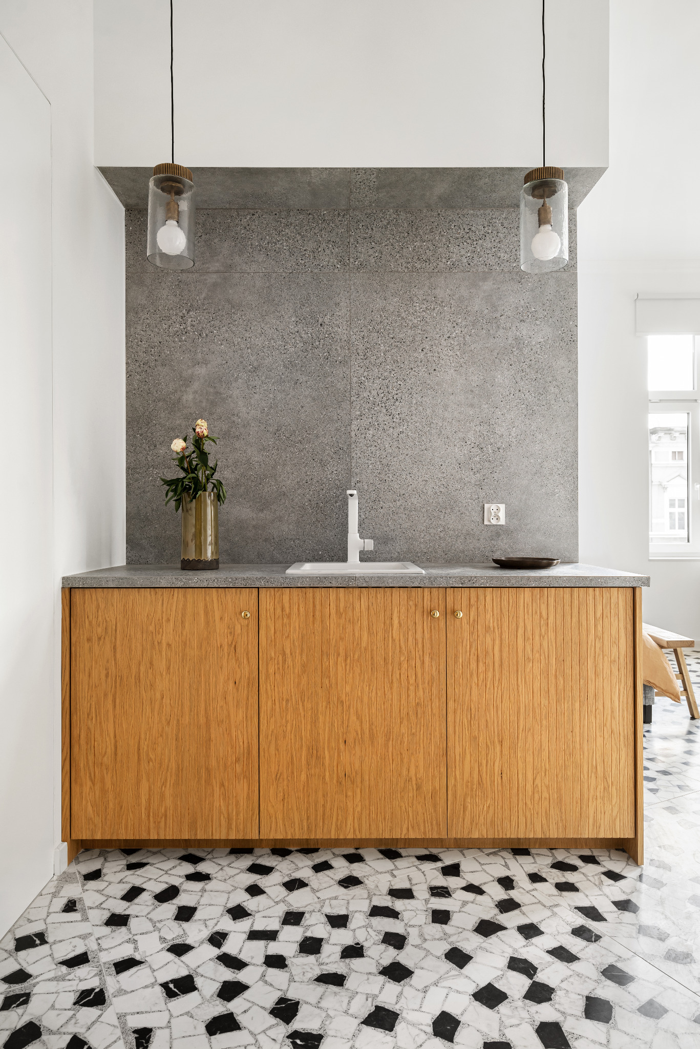 Nowoczesna łazienka z drewnianą szafką pod umywalką, szarym blatem i mozaikową podłogą w czarno-białe wzory. Na blacie wazon z kwiatami i czarna miseczka. Dwie szklane lampy wiszą nad blatem.