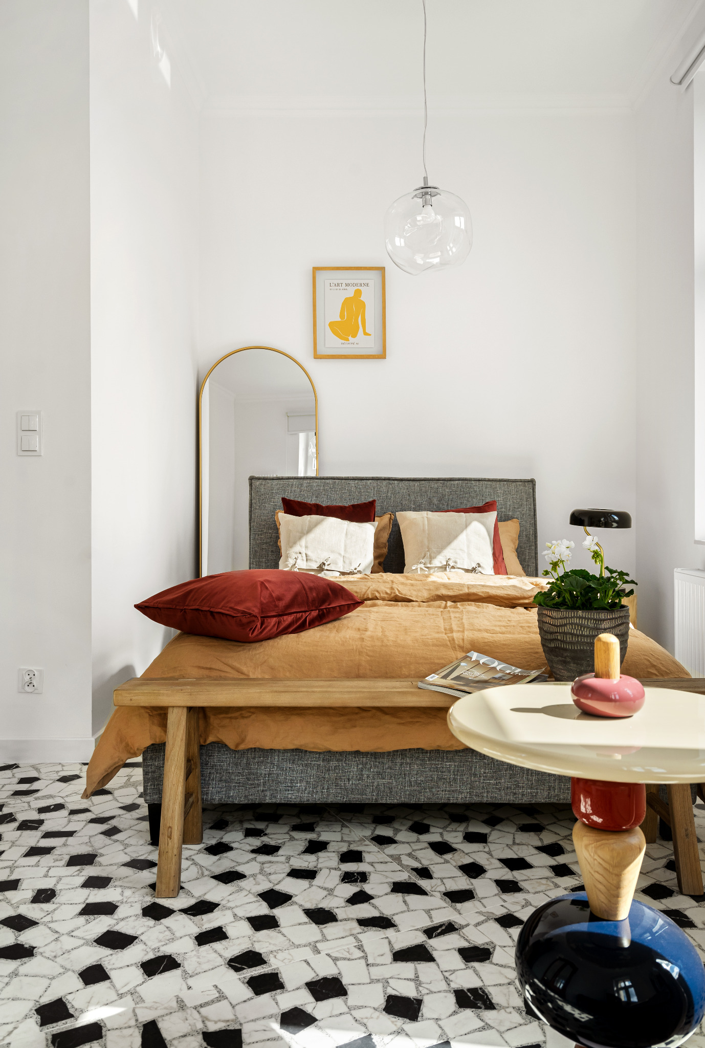 Jasna sypialnia z łóżkiem z szarą ramą i drewnianym wezgłowiem, nakryta brązową pościelą i burgundowymi poduszkami, uzupełniona o złote lustro, modernistyczny stolik boczny i podłogę w czarno-białą...