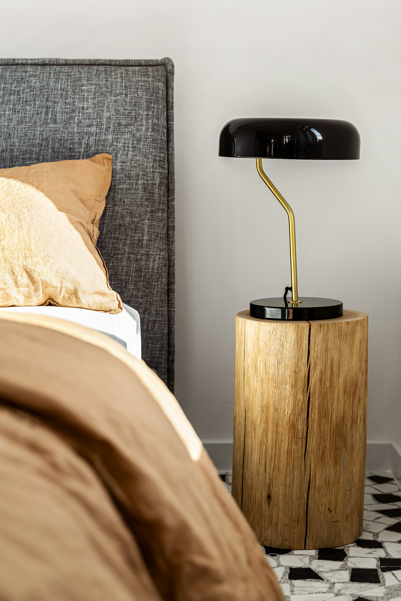 Sypialnia z łóżkiem z szarym wezgłowiem, złotą pościelą i lampką nocną z czarnym kloszem stojącą na drewnianym pniu jako stoliku nocnym, na podłodze biało-czarna mozaika.