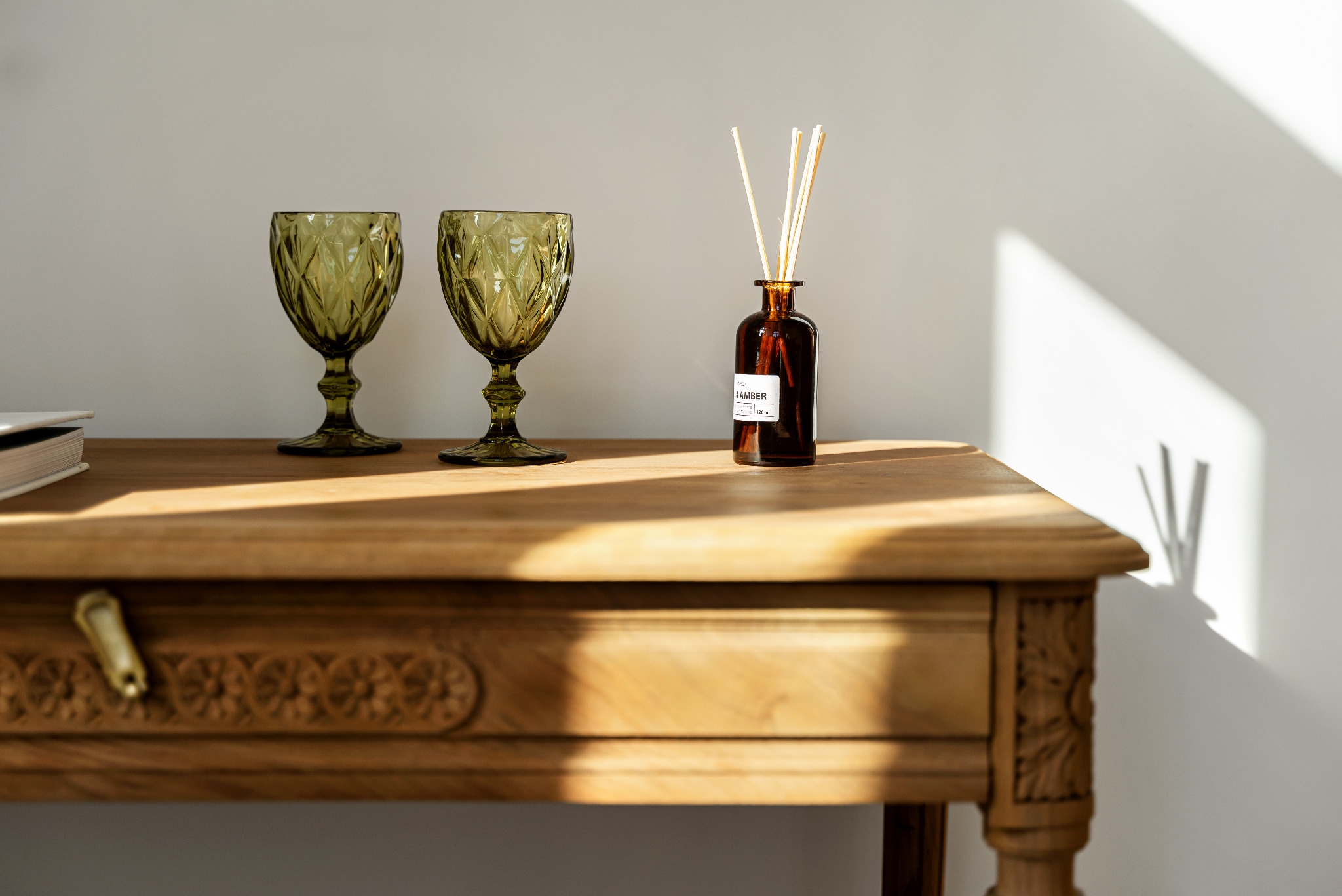 Dwa zielone kieliszki i brązowy dyfuzor zapachowy z patyczkami na zdobionym, drewnianym stoliku w jasnym świetle.