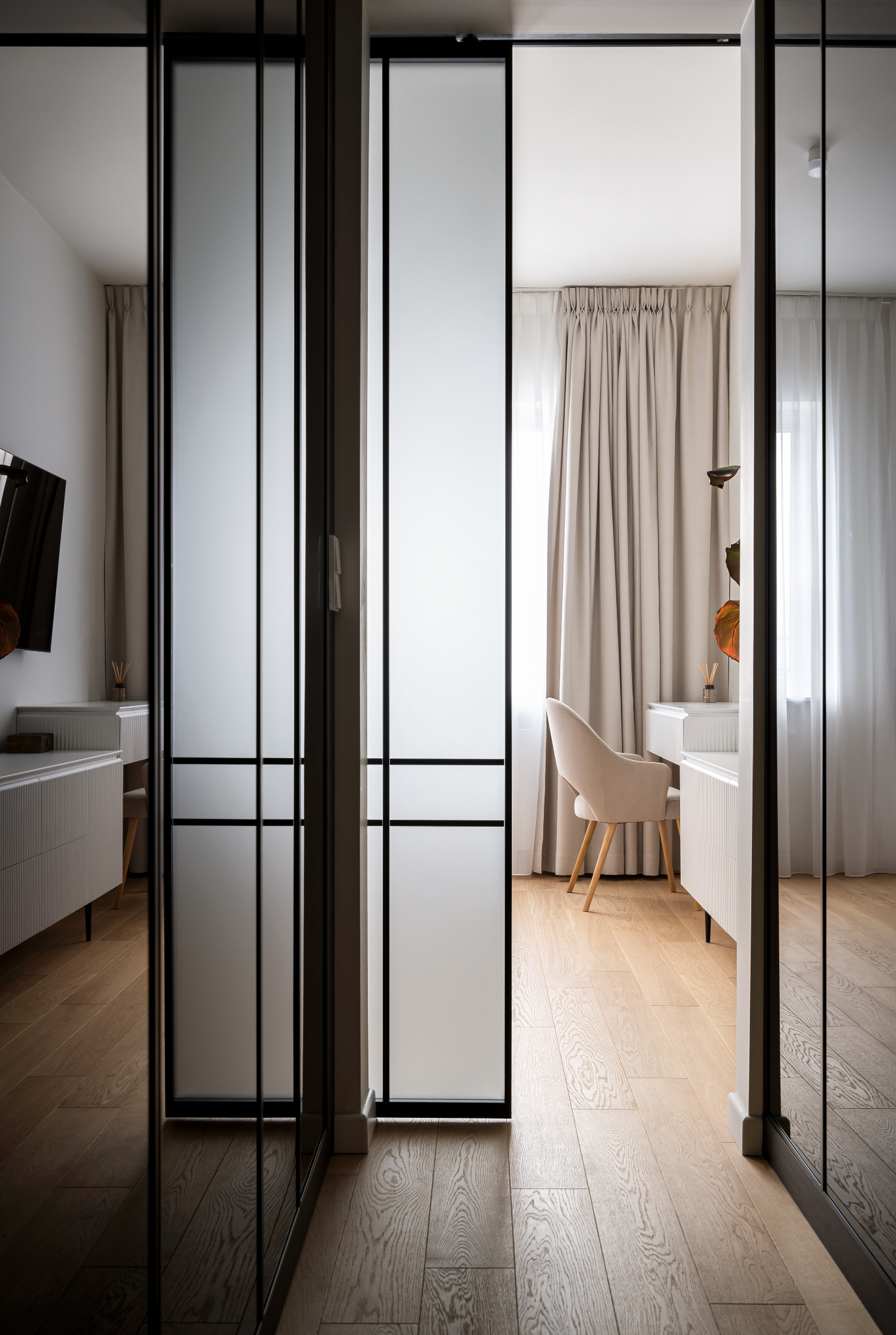 Nowoczesne wnętrze mieszkania z przesuwnymi drzwiami ze szkła matowego w czarnej ramie, widok na toaletkę z beżowym krzesłem i zasłony w neutralnych kolorach.