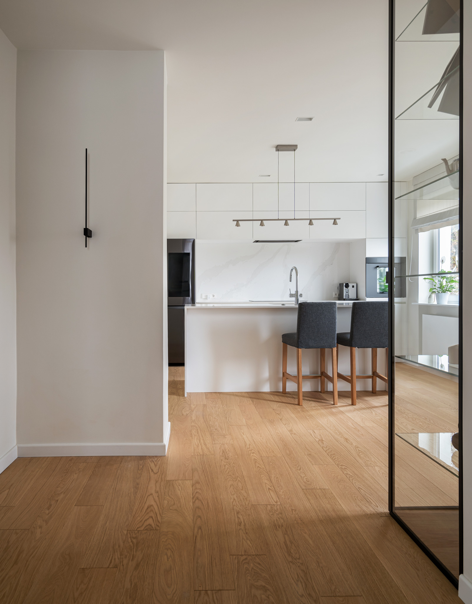 Nowoczesna, minimalistyczna kuchnia z wyspą, dwoma szarymi hokerami, białymi szafkami i drewnianą podłogą, widoczna z perspektywy przedpokoju.