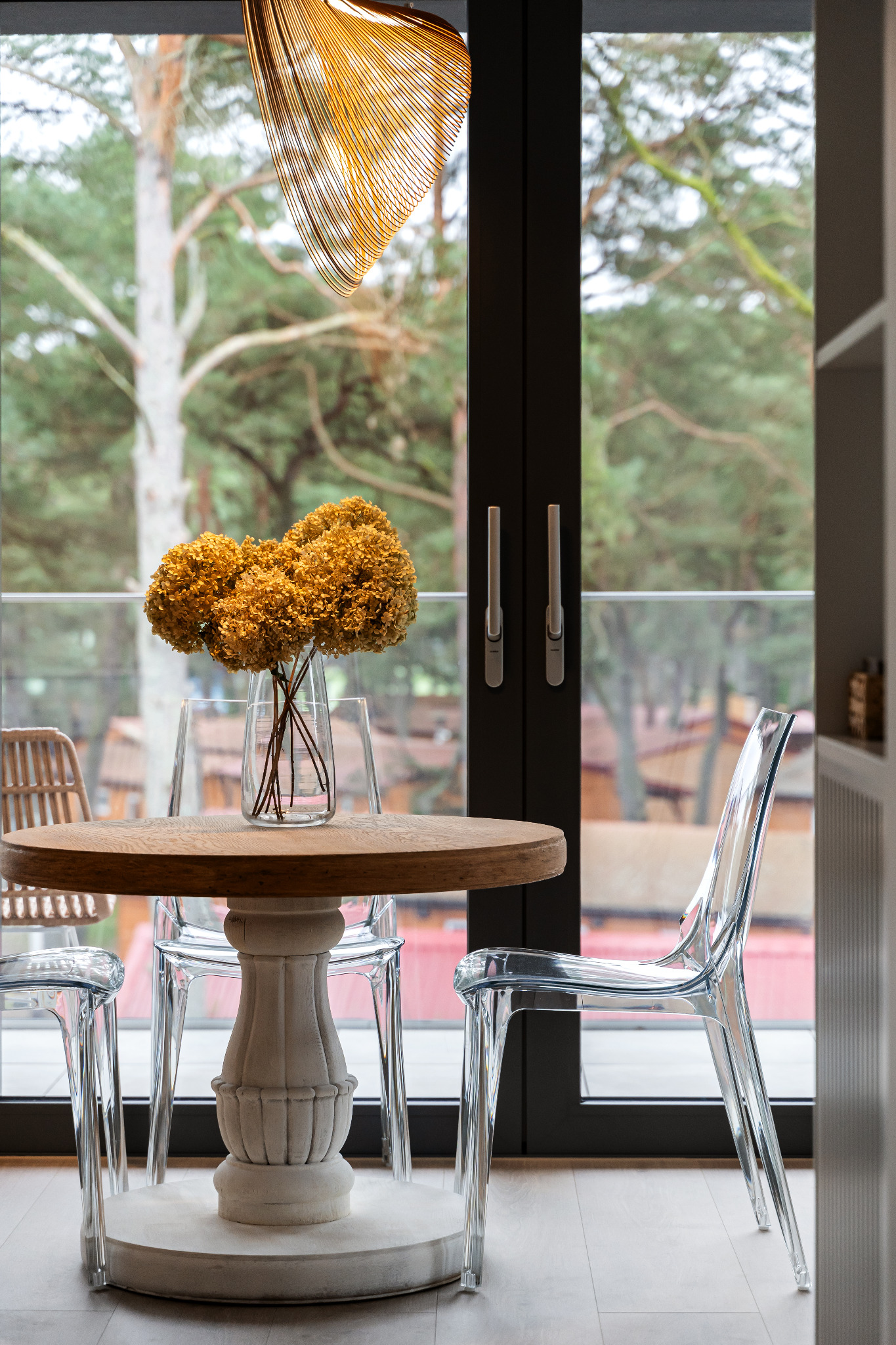 Okrągły drewniany stół z dekoracyjnym bukietem suszonych hortensji w szklanym wazonie, otoczony przezroczystymi krzesłami, na tle okna z widokiem na las.