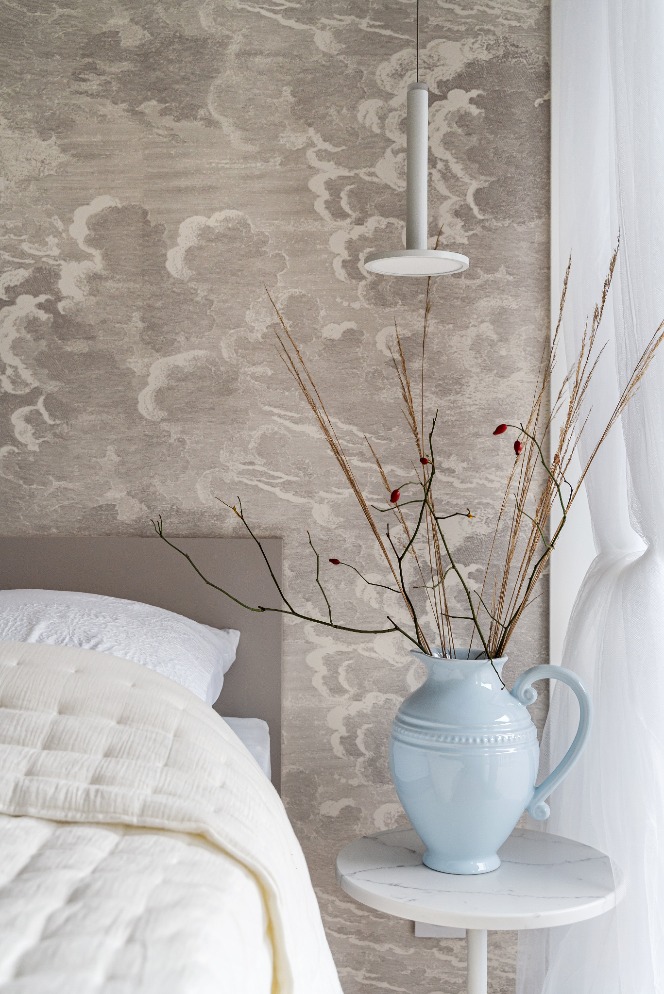 Sypialnia z łóżkiem przykrytym białą narzutą, stolik nocny z marmurowym blatem, na którym stoi jasnoniebieski wazon z suchymi trawami i gałązkami z czerwonymi owocami, w tle tapeta w chmury...