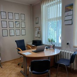 Biuro Ubezpieczeń Inwesta Ewa Wieczorek-Syjut - Biuro z biurkiem, laptopem i krzesłami. Na ścianie certyfikaty i kalendarz. Widok na okno z żaluzjami pionowymi. Stonowana kolorystyka wnętrza.