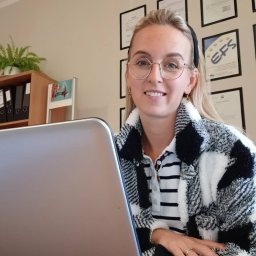 Biuro Ubezpieczeń Inwesta Ewa Wieczorek-Syjut - Uśmiechnięta blondynka w okularach, siedząca za laptopem w biurze z certyfikatami na ścianie. Widoczny fragment laptopa i dłoń na klawiaturze.