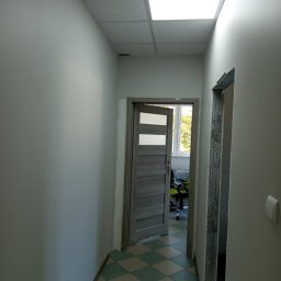 Krzysztof Kleban - Korytarz po remoncie: szare ściany, podłoga w zielono-białą kratę, dwoje drzwi, jedne otwarte z widokiem na biuro, drugie w trakcie montażu ościeżnicy.