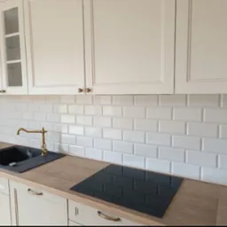 Biała kuchnia z płytkami typu metro, złotą armaturą i drewnianym blatem. Widoczne szafki górne z przeszklonymi frontami i płyta indukcyjna.