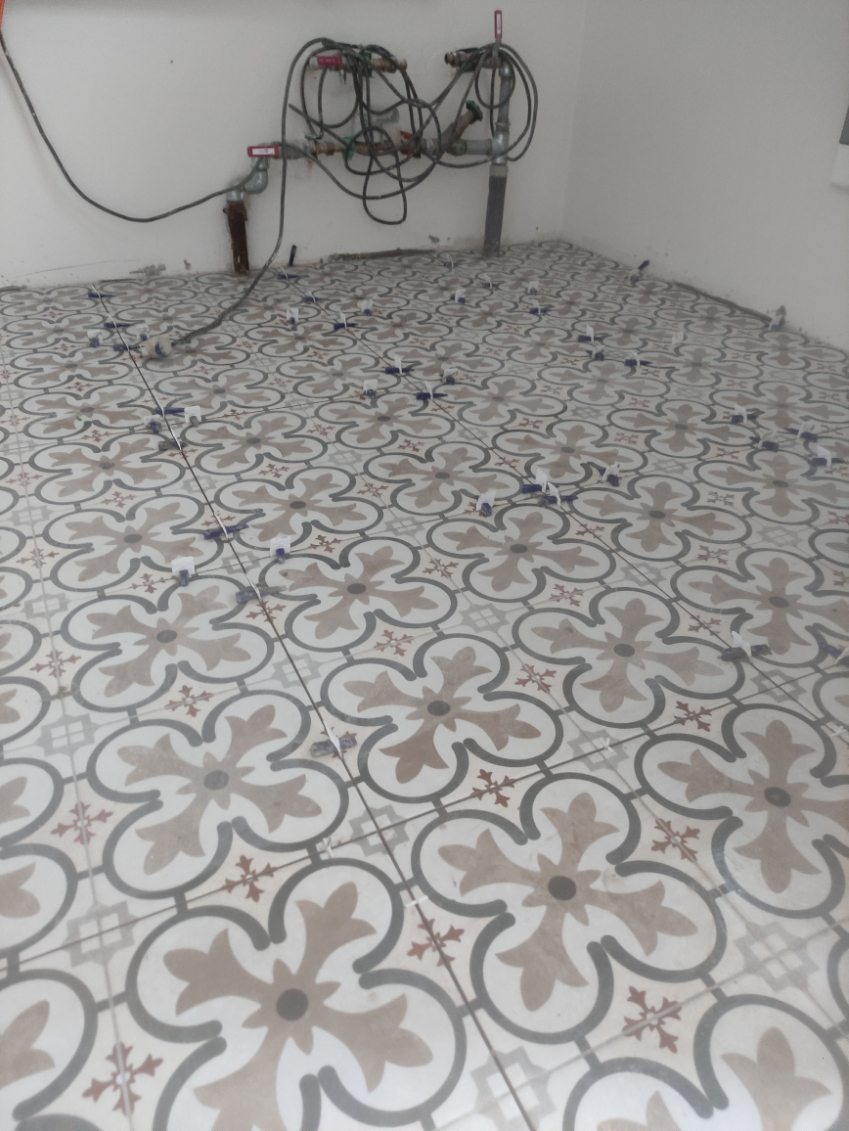 Układanie dekoracyjnych płytek podłogowych w łazience z krzyżykami dystansowymi, widoczne rury instalacyjne przy ścianie. Wzór kwiatowy na płytkach.