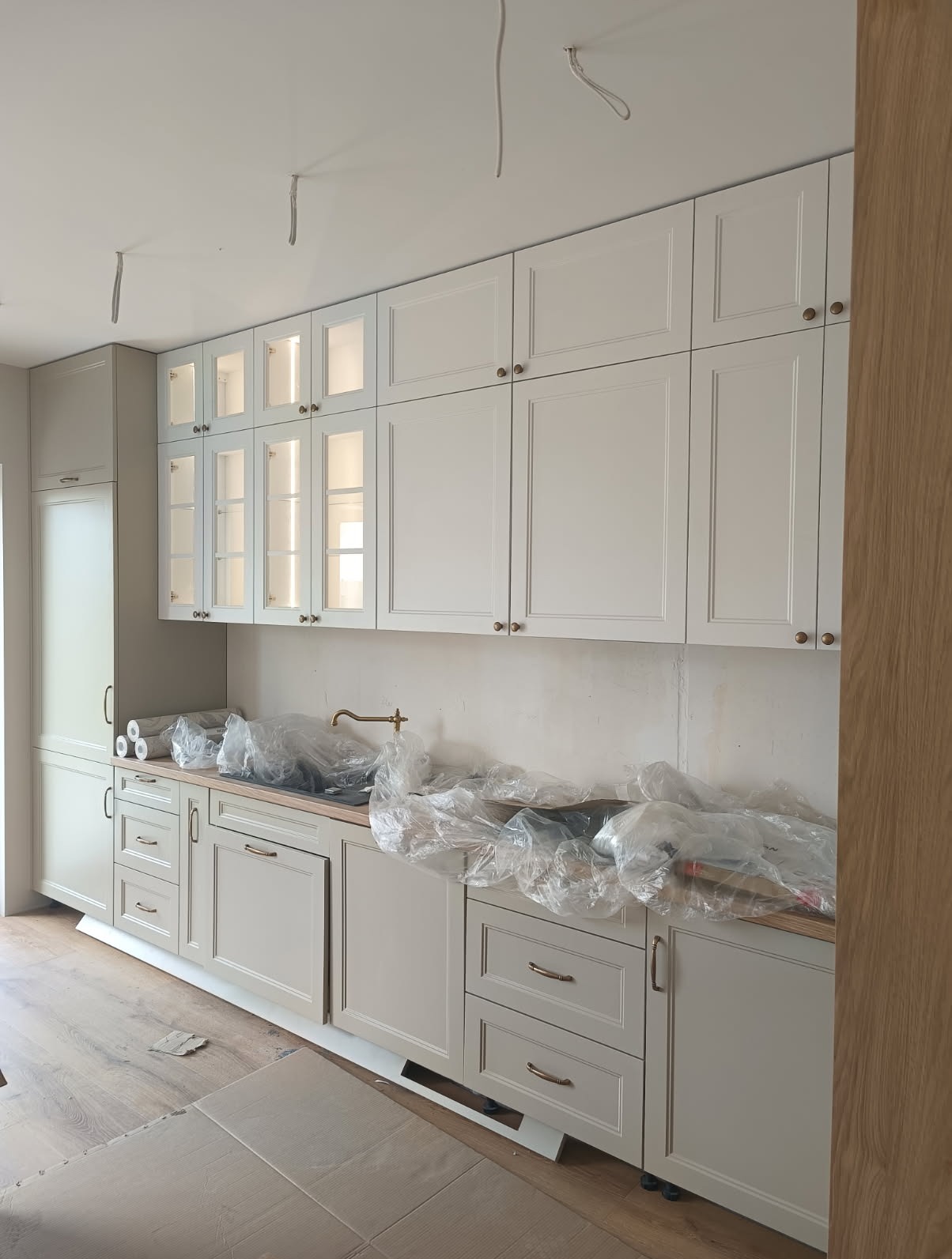 Jasna kuchnia z kremowymi szafkami w stylu klasycznym, częściowo zakryta folią ochronną, z widoczną złotą baterią i niedokończonymi elementami wykończenia.