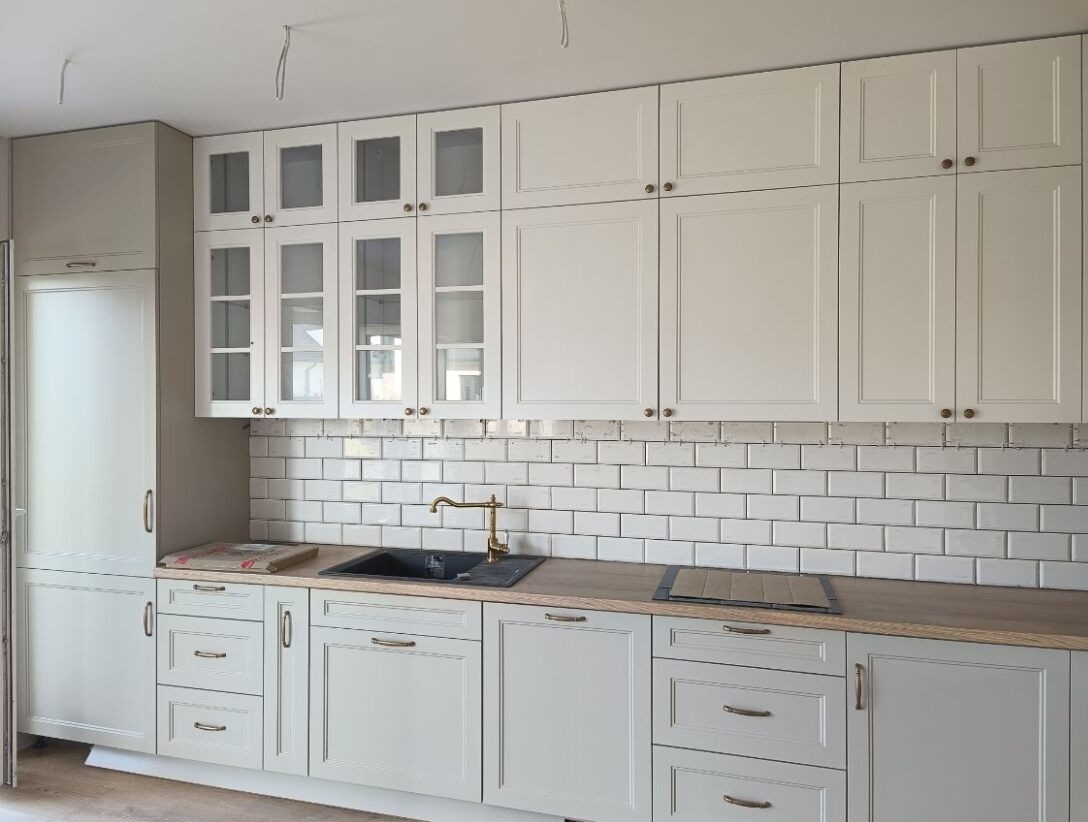 Jasna kuchnia w stylu retro z białymi płytkami i drewnianym blatem, złota bateria. Widoczne górne szafki z przeszkleniami i dolne szafki z szufladami.