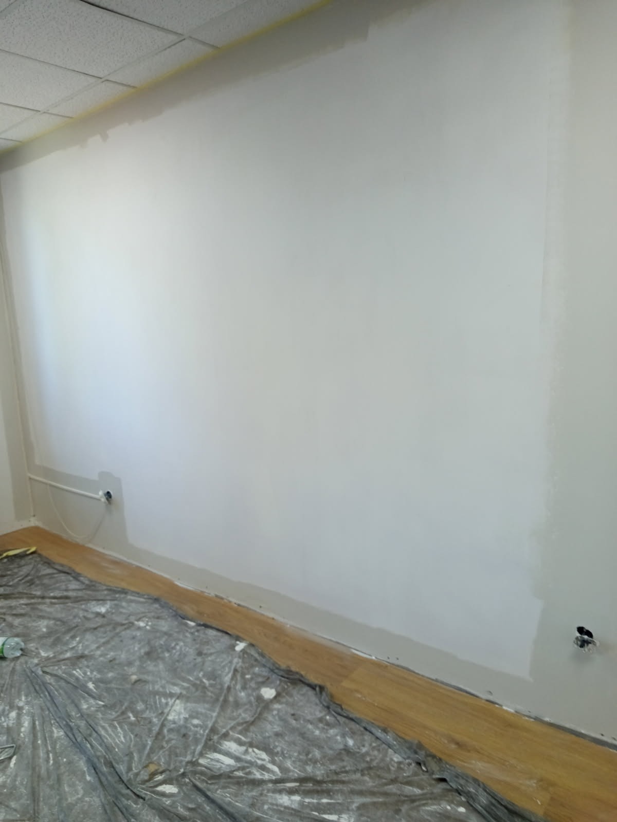 Ściana w trakcie malowania na biało, zabezpieczona folią podłoga. Widoczne ślady farby na folii i częściowo pomalowana ściana w pomieszczeniu.